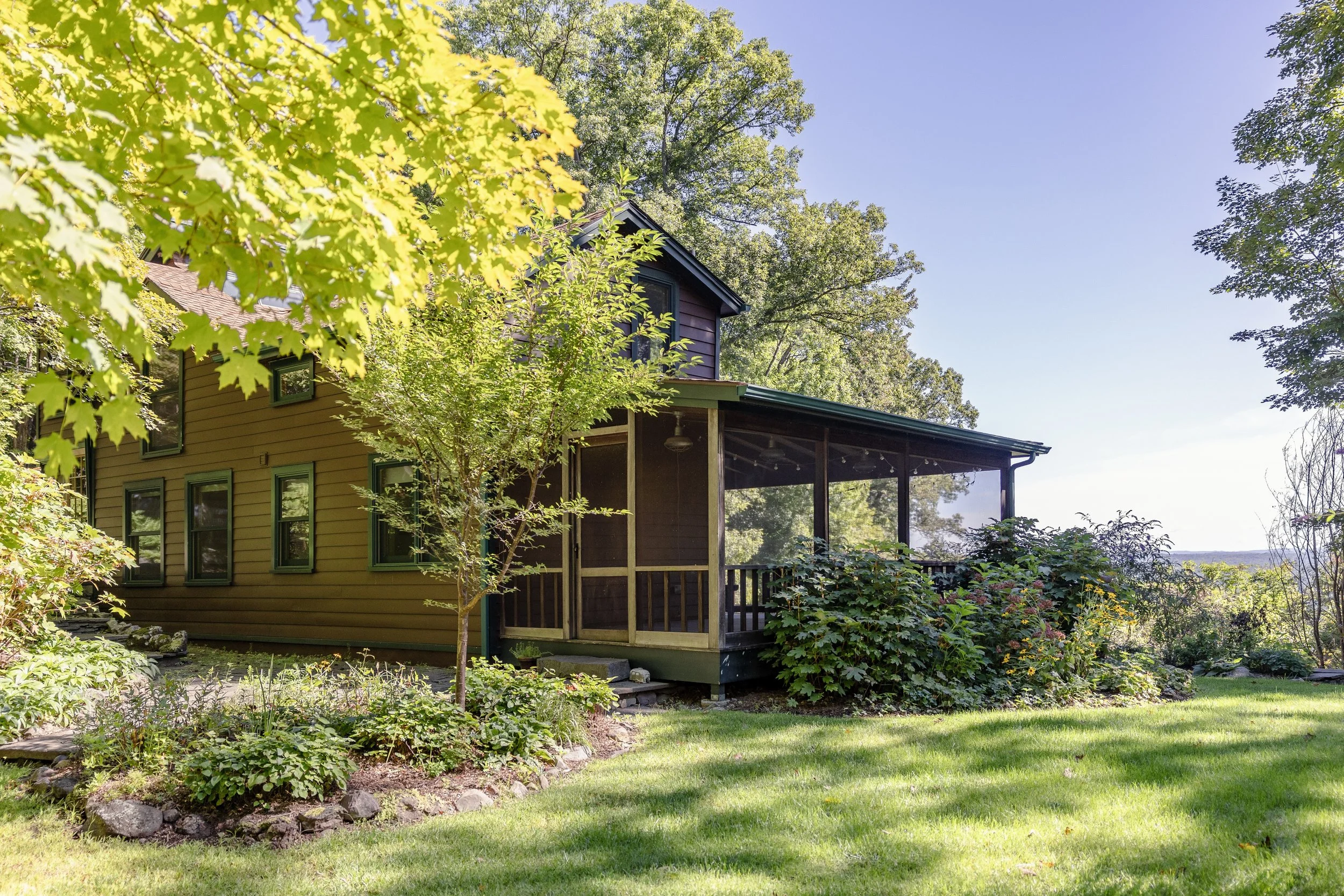 A house with green and purple exterior, surrounded by lush trees and a well-maintained lawn, with a screened porch on the side.