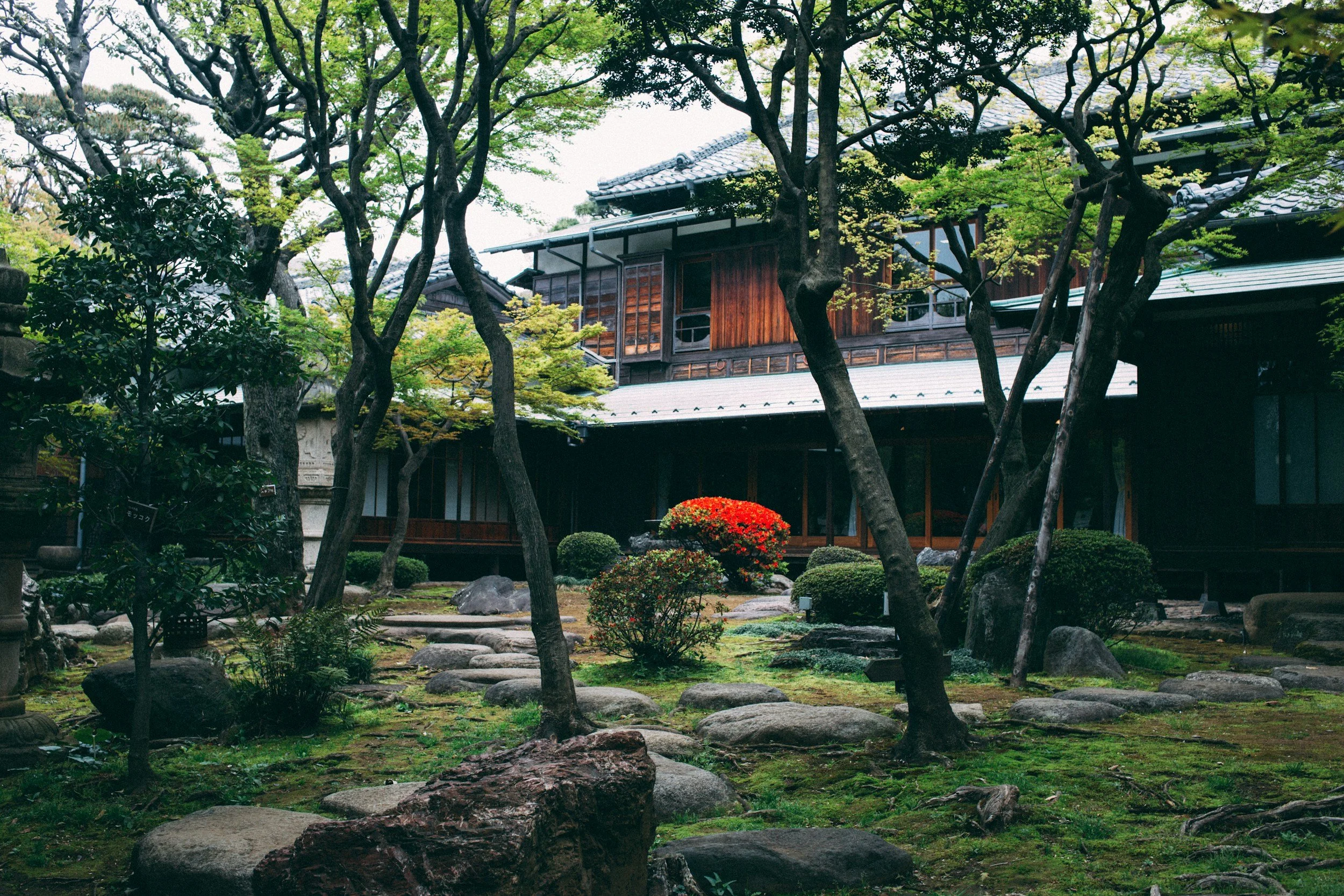 Traditional Japanese garden with trees, rocks, and a wooden building in the background.