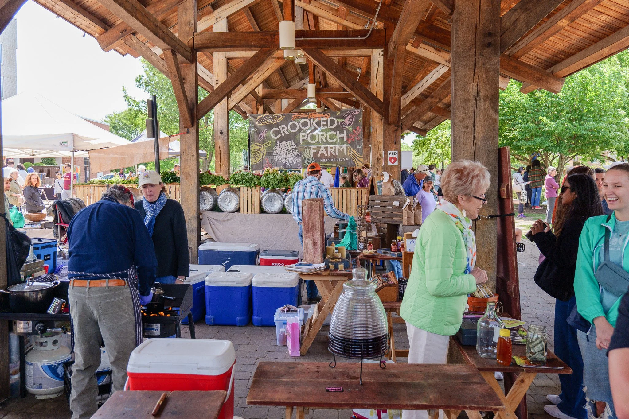 People shopping at an outdoor farmer's market under a wooden pavilion on a sunny day, with a sign reading 'Crooked Porch Farm' in the background.