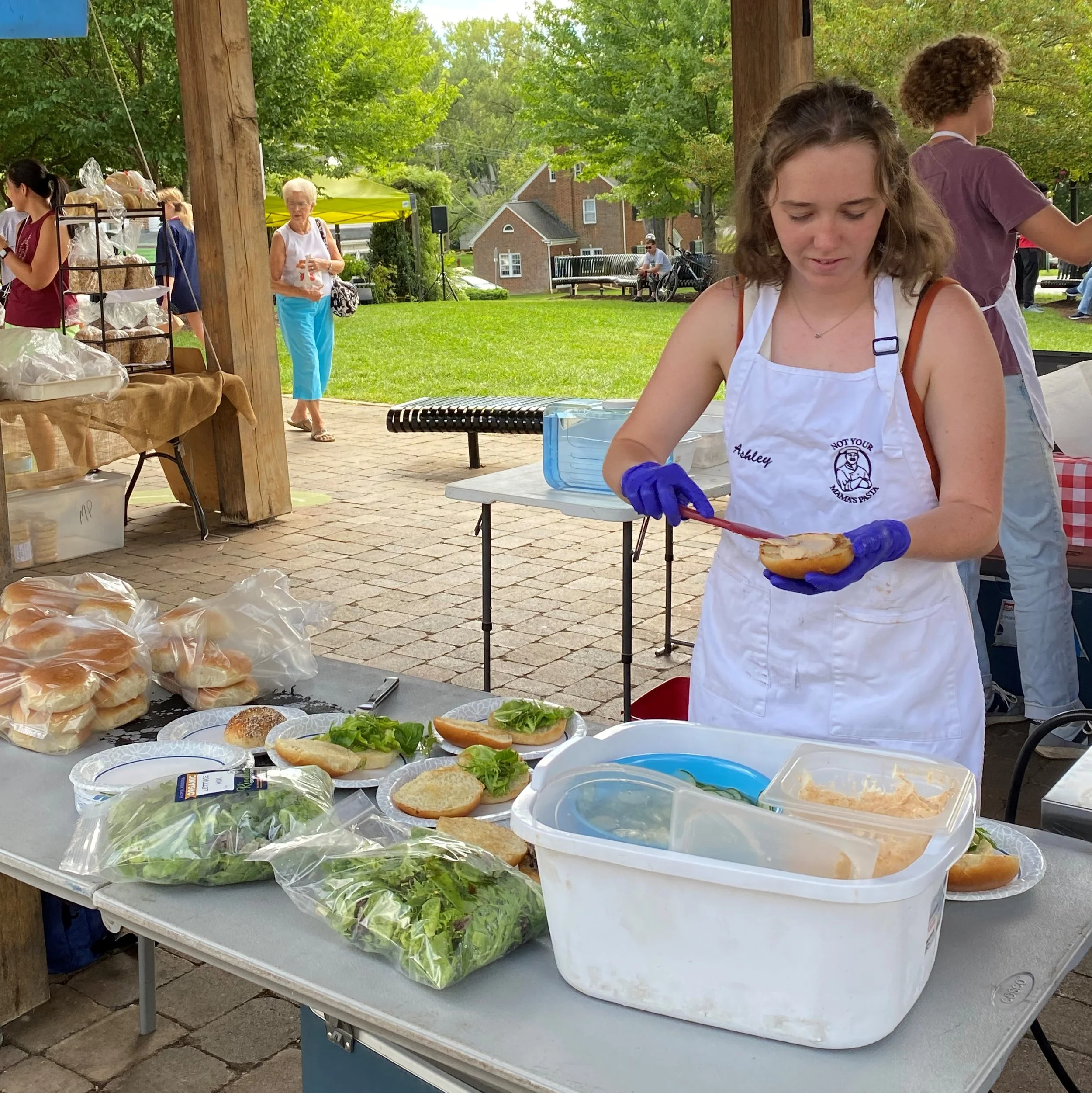 A young woman wearing a white apron and purple gloves is preparing a sandwich at an outdoor food booth with buns, lettuce, and condiments on the table. In the background, there are people walking in a park-like setting with green trees, benches, and a small yellow tent.