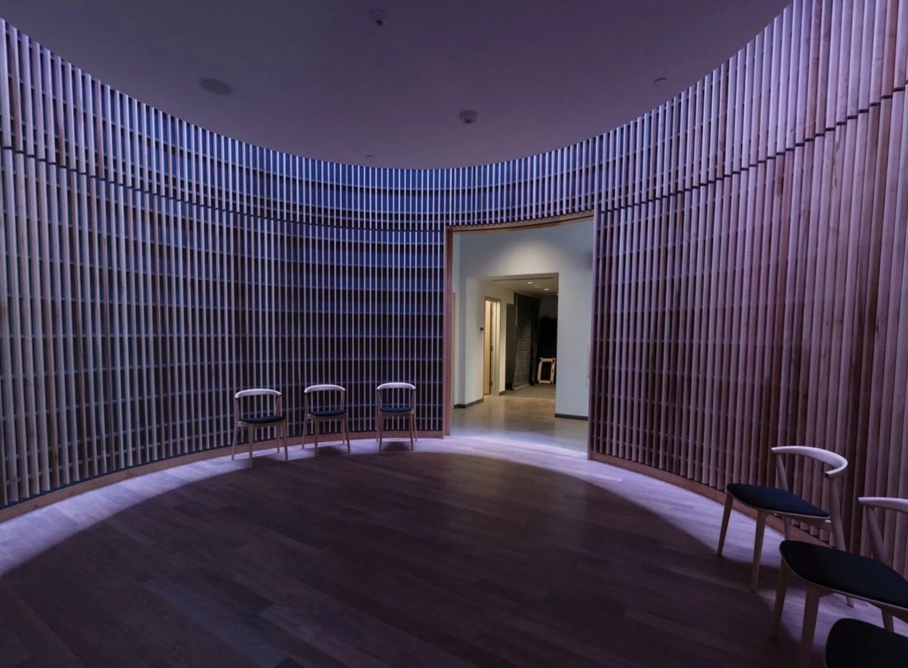 Empty room with wooden slat walls, curved design, four white chairs with black seats along the wall, and a doorway leading to a dimly lit hallway.