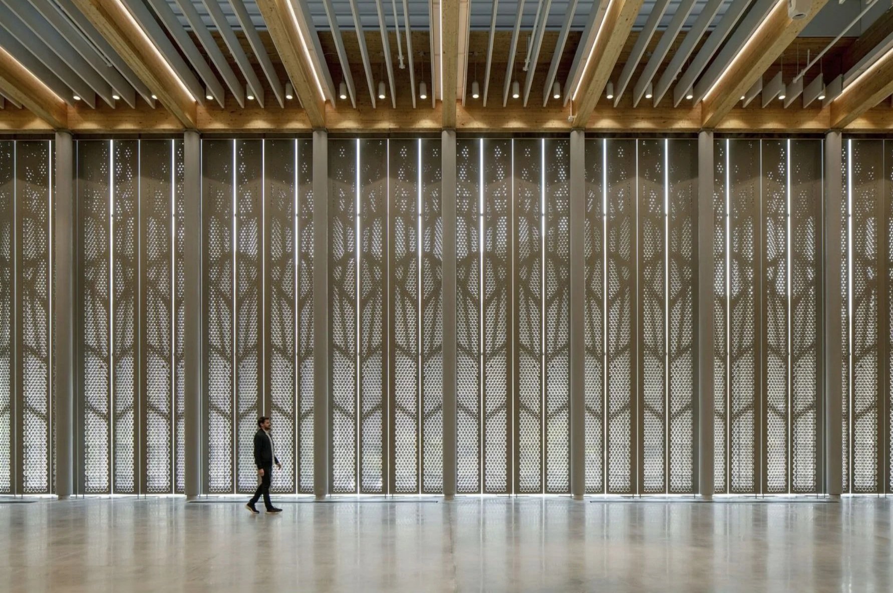 A man in a black suit walking in a spacious, modern interior with large decorative metal panels casting intricate shadows, wooden beams on the ceiling, and a light-colored floor.