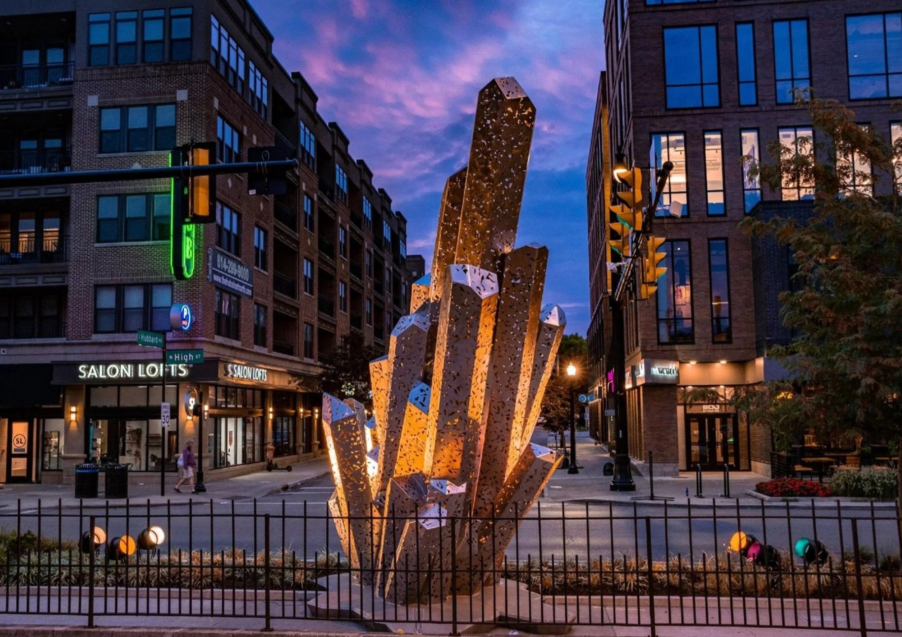 A modern downtown street scene at dusk with a large illuminated abstract sculpture in the center. Surrounding buildings are made of brick and glass, with some storefronts visible, including 'Salon Lofts.' The sky is purple and pink with clouds, and s