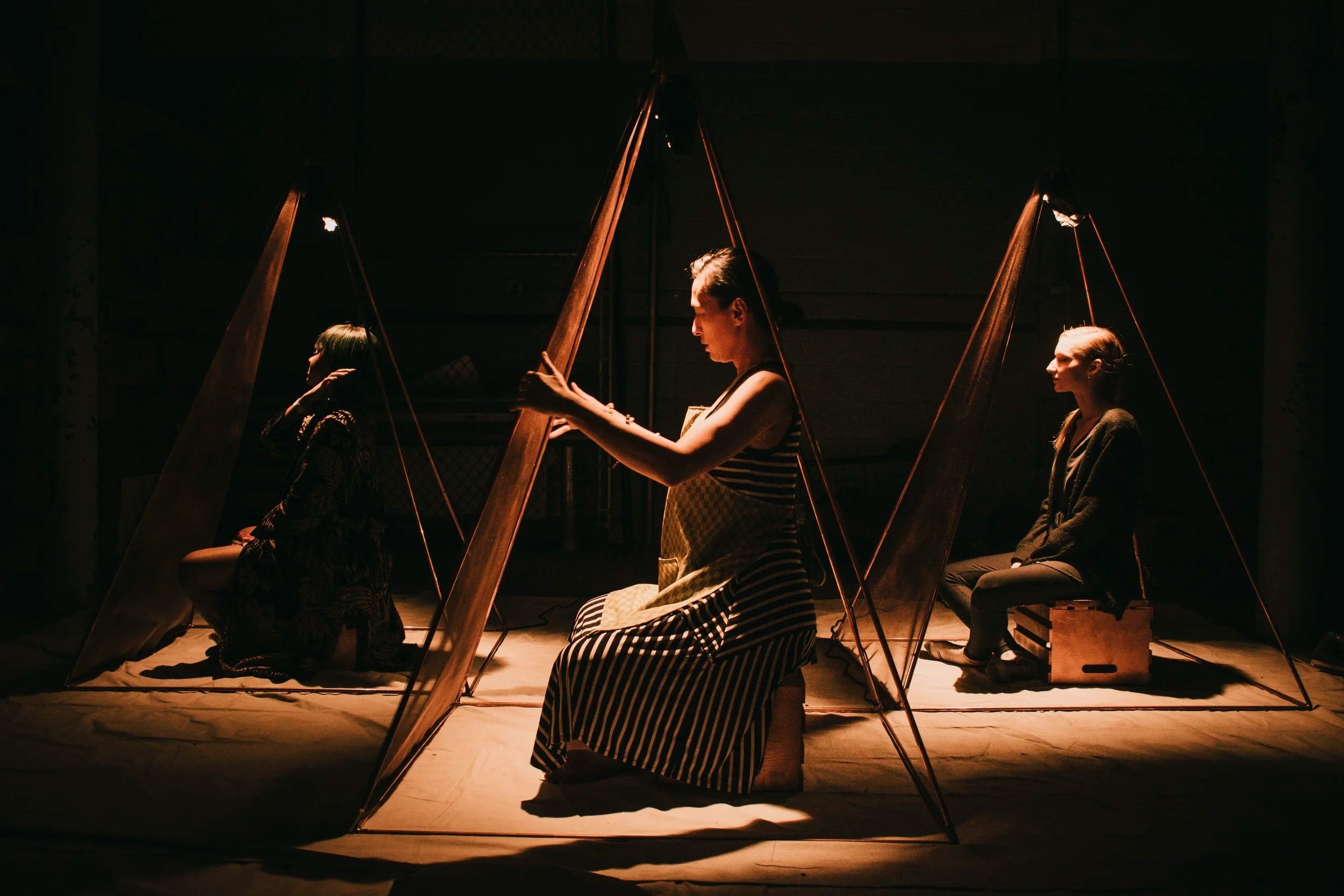 Three women seated inside fabric tents illuminated by warm lighting, engaged in contemplative or meditative activities.