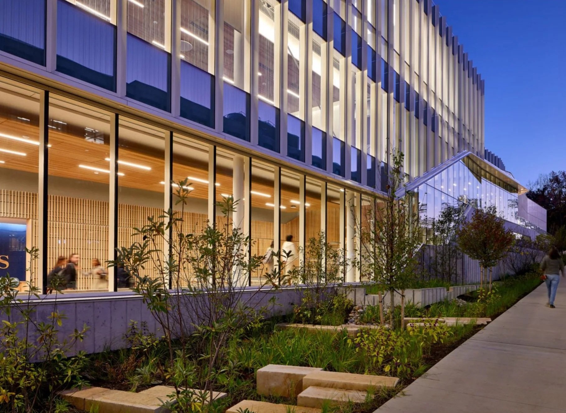 Modern multi-story building with glass exterior and warm interior lighting, surrounded by landscaped greenery and a concrete walkway at dusk.
