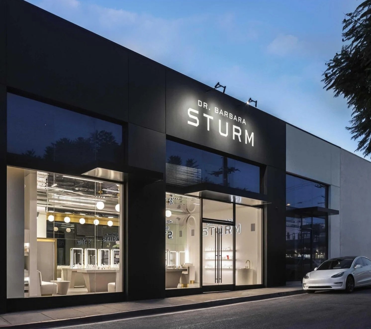 Modern storefront of Dr. Barbara Sturm with large glass windows and interior lighting, featuring a white car parked outside during the evening.