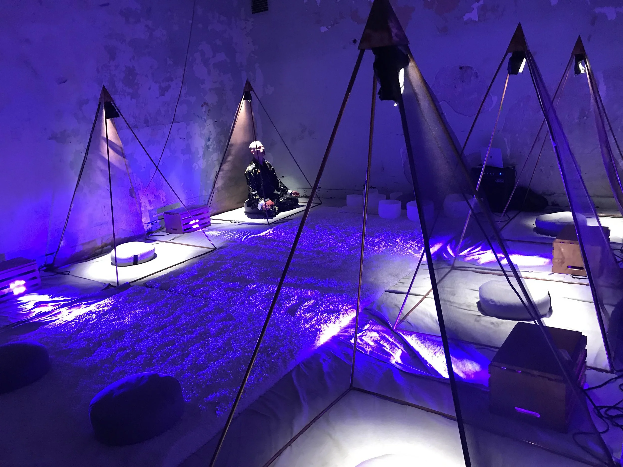 A person in dark clothing sitting in a meditation pose within a black pyramid-shaped tent illuminated by purple and white lights, with cushions and a textured surface inside.