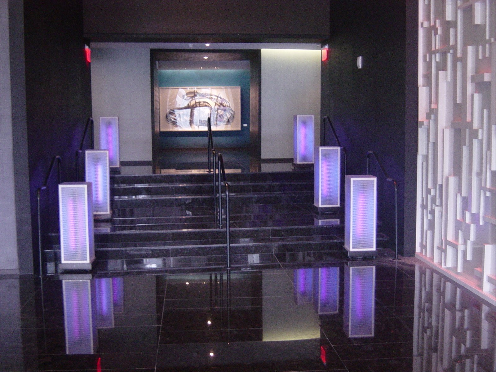 Modern hotel lobby entrance with black marble stairs, purple lighting accents on white pillars, and an abstract artwork on the wall at the top of the stairs.