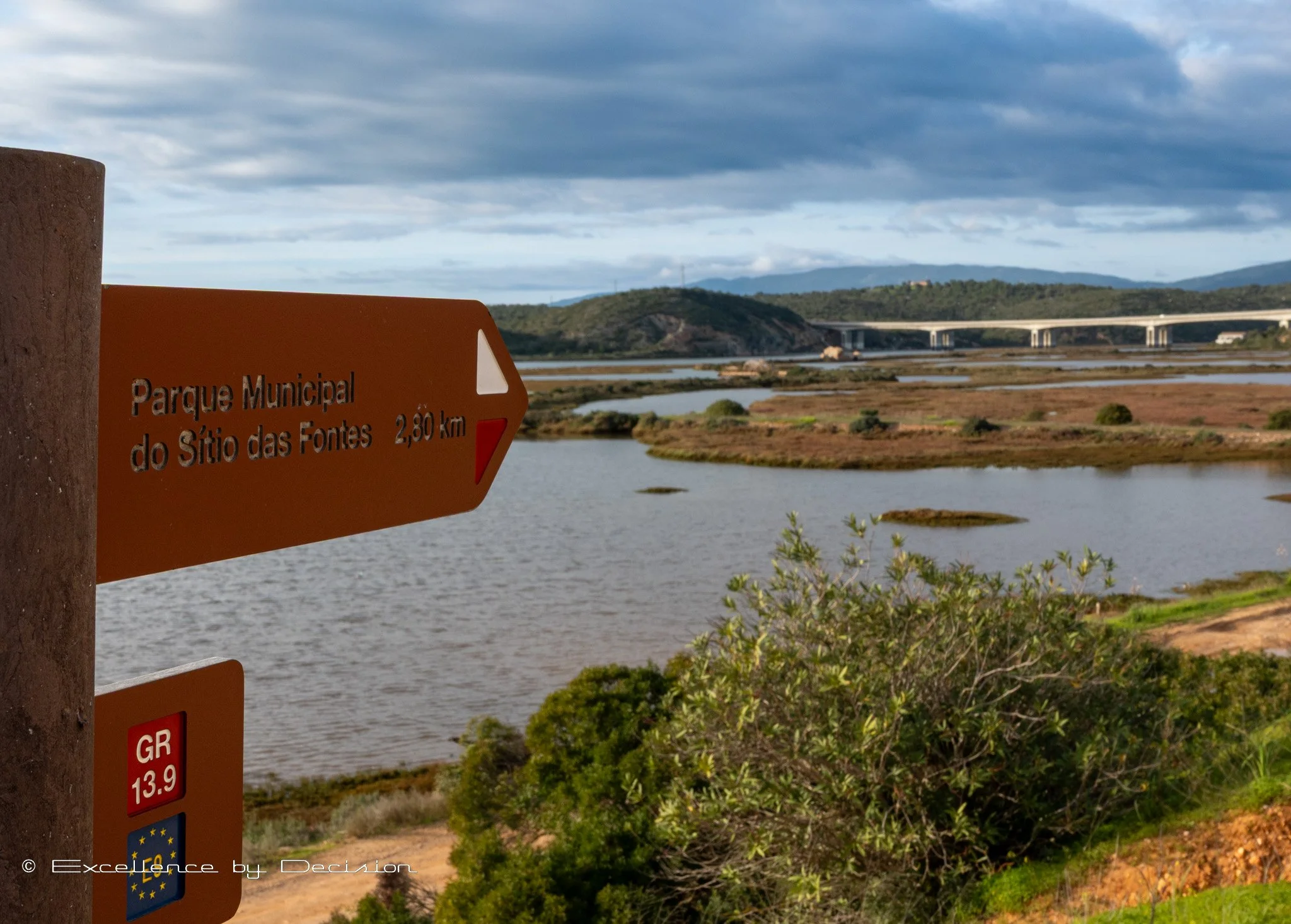 Sign for Parque Municipal do SÃtio das Fontes, 2.8 km away, with landscape and bridge in background.