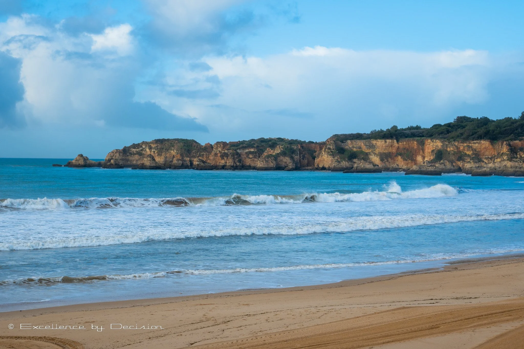 Scenic view of a sandy beach with ocean waves and rocky cliffs in the background under a blue sky.
