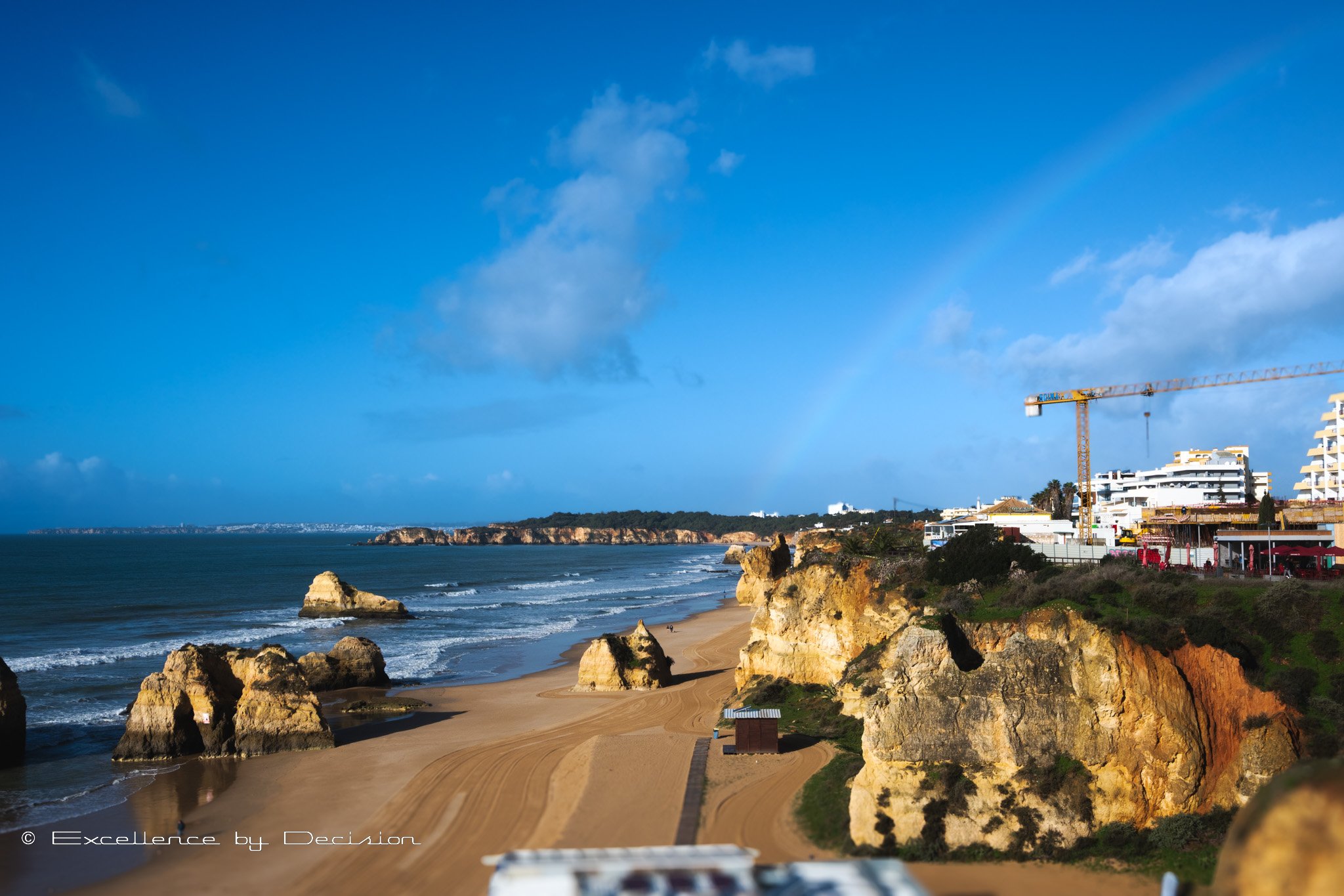 Beach with rocky cliffs, waves, a crane, buildings, and a rainbow in the sky.