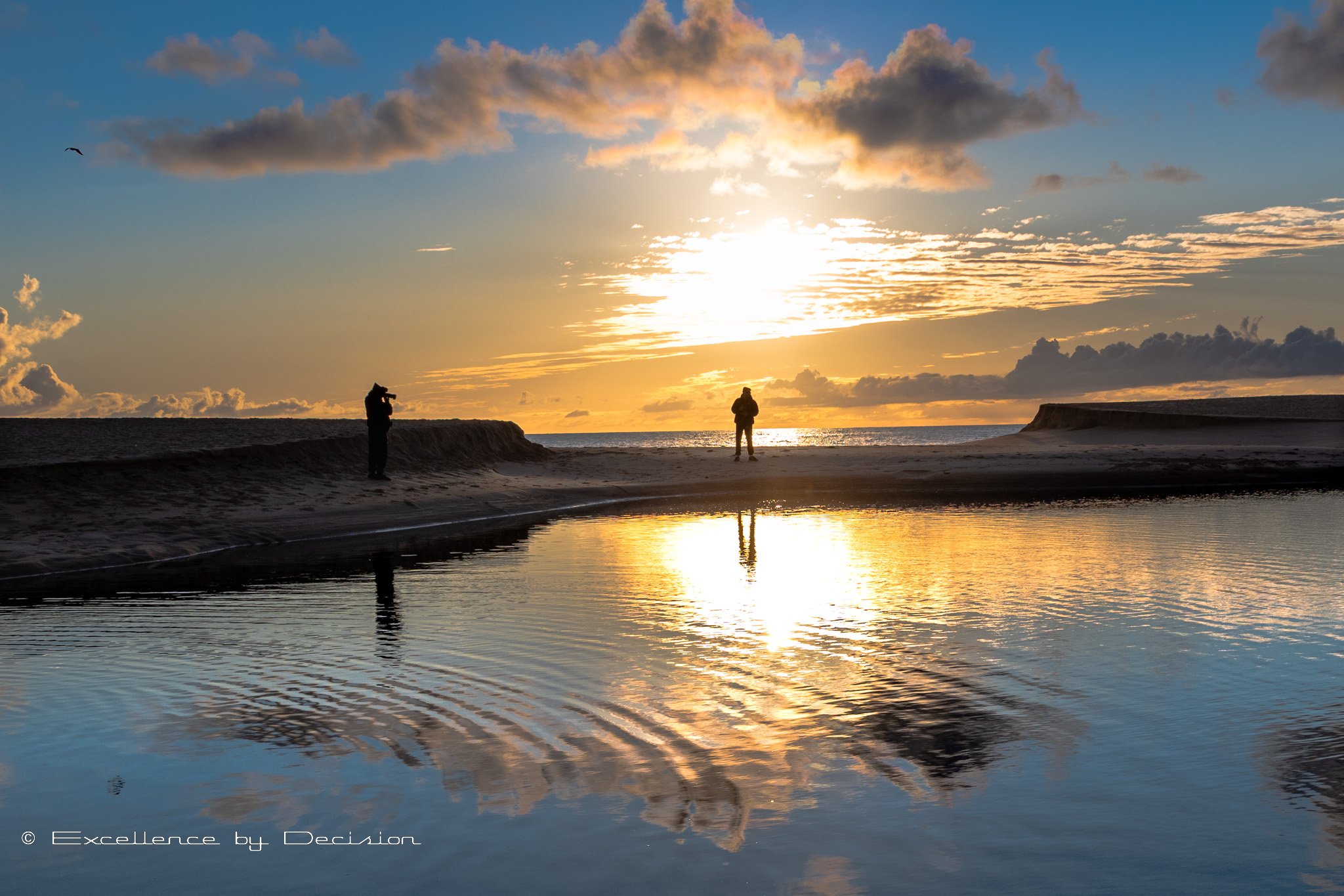 Beach sunset with two silhouetted photographers, tranquil water reflecting sky and clouds, bird flying, and vibrant orange and blue sky.