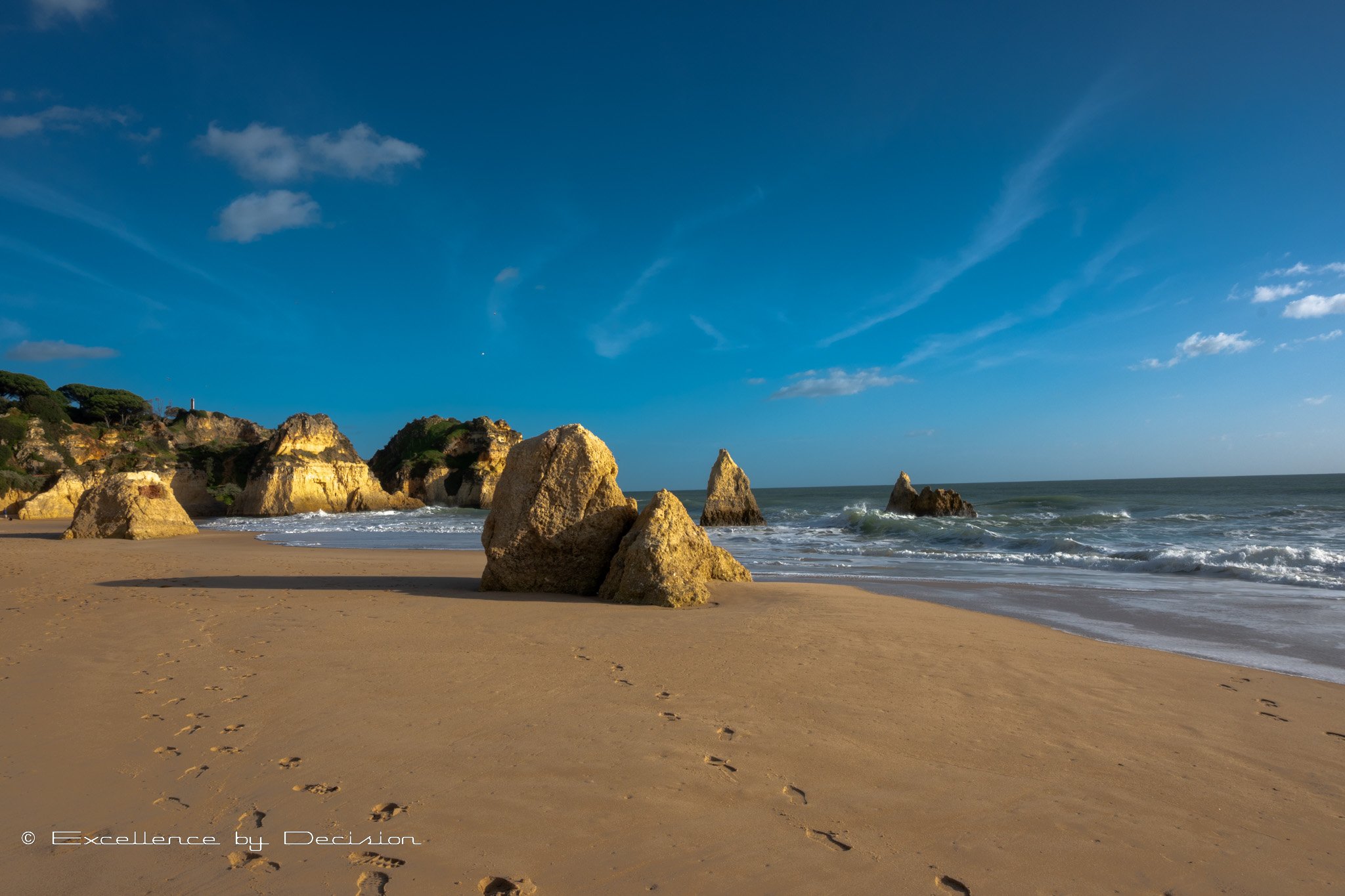 Rocky beach with golden sand, ocean waves, and clear blue sky. Large rocks scattered across the shoreline.