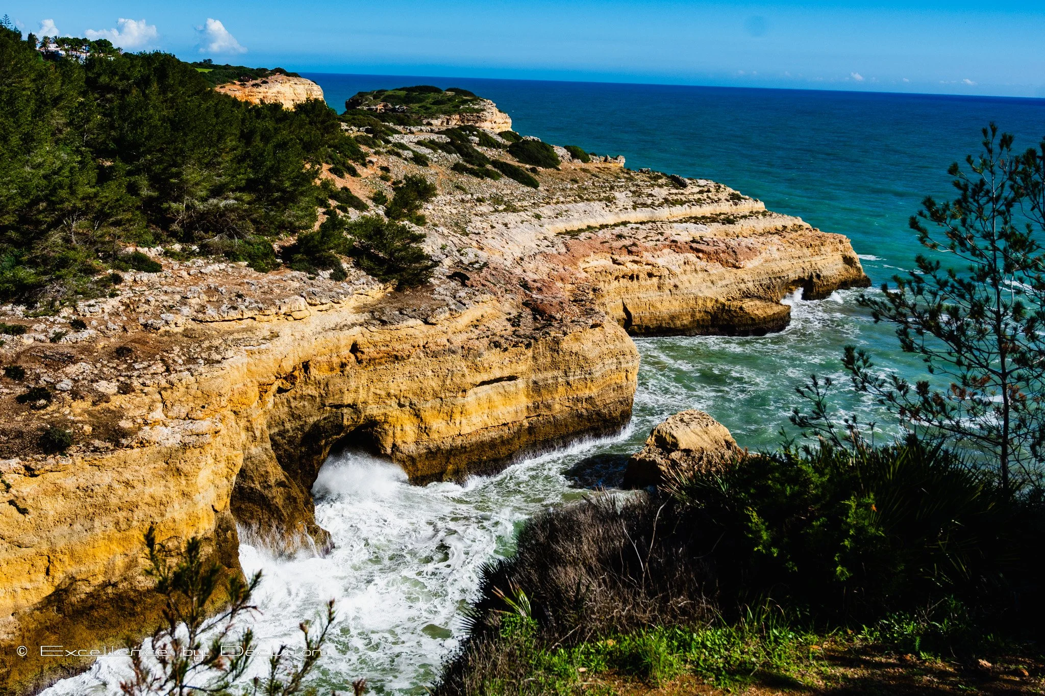 Rocky coastal cliffs with clear blue ocean, green vegetation, and sea caves, Algarve, Portugal.
