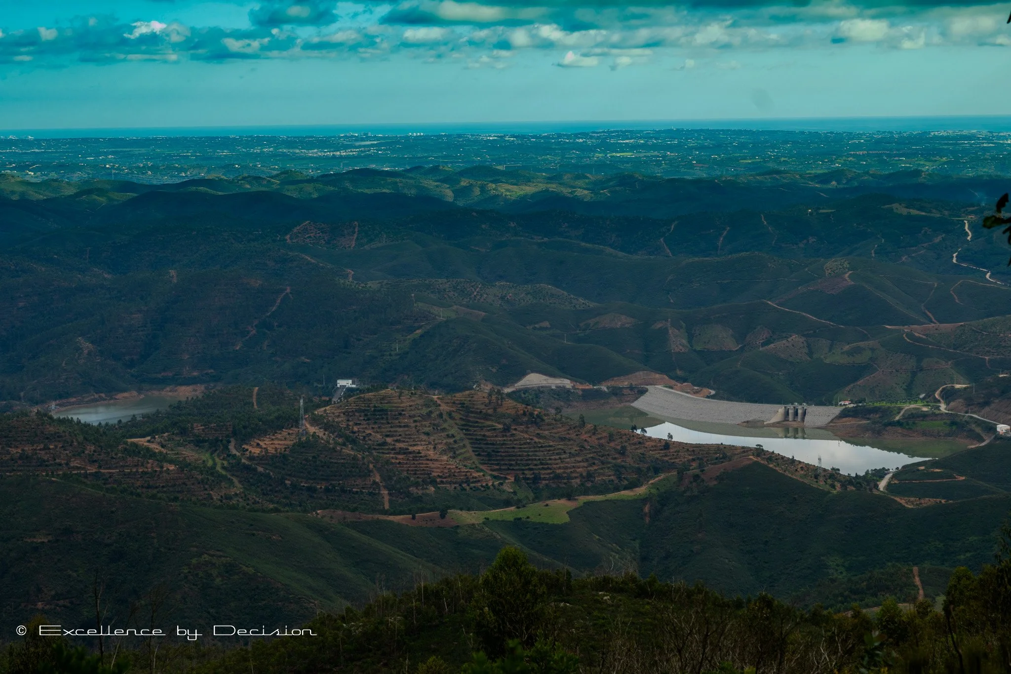 Aerial view of rolling green hills with a small reservoir and dam in the foreground, under a partly cloudy sky.