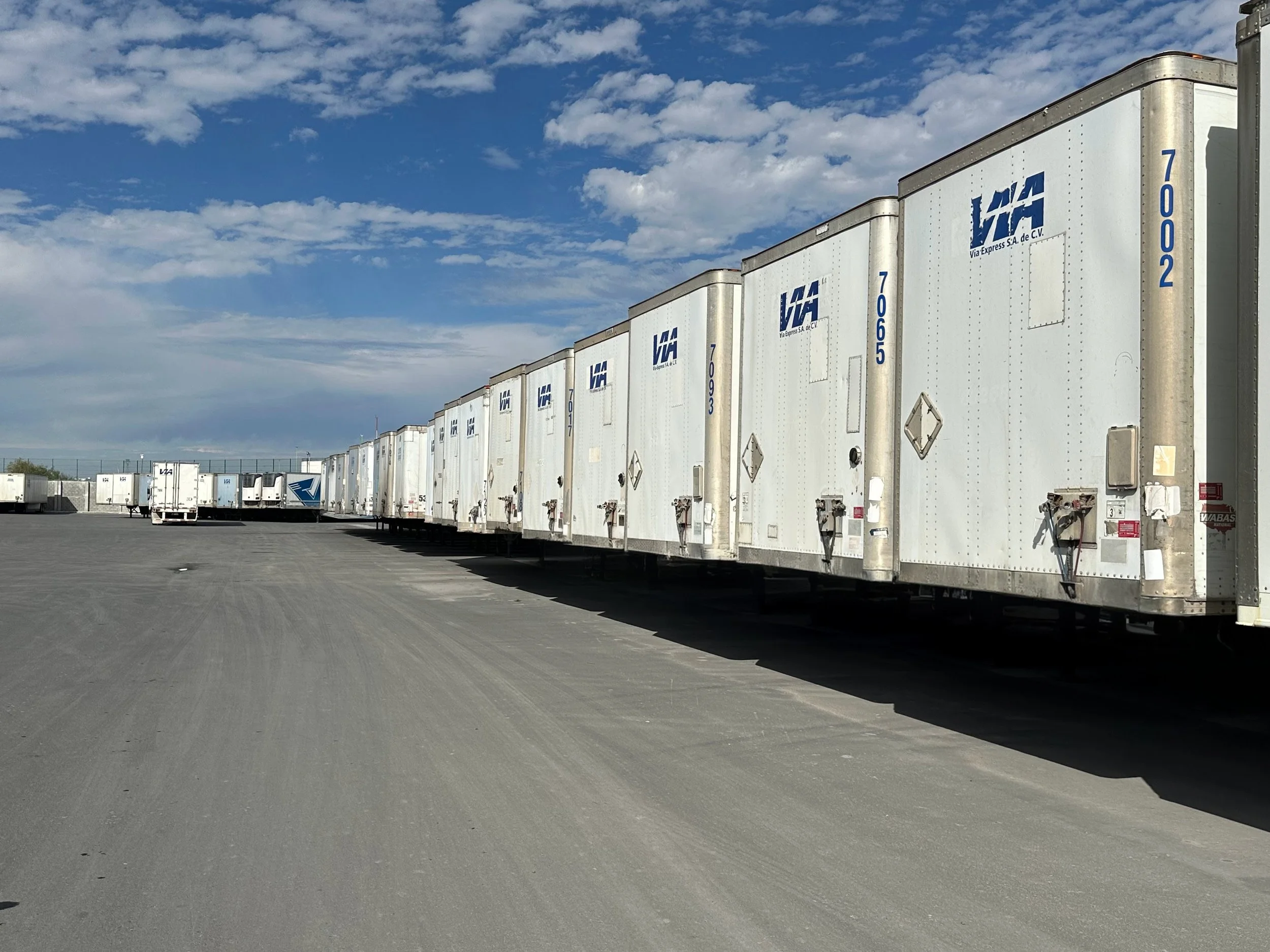 A row of white semi-truck trailers parked in an open lot under a partly cloudy sky.