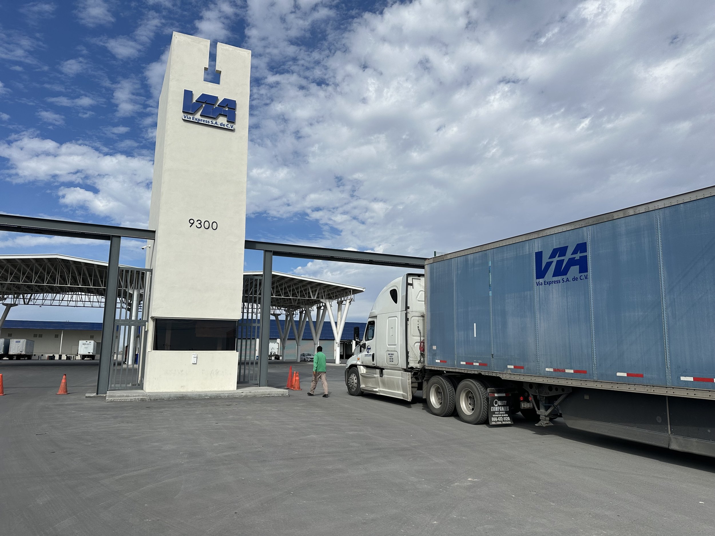 A parking lot with a large white sign displaying the LAS logo and address, alongside trucks and a man walking. The sky is partly cloudy.
