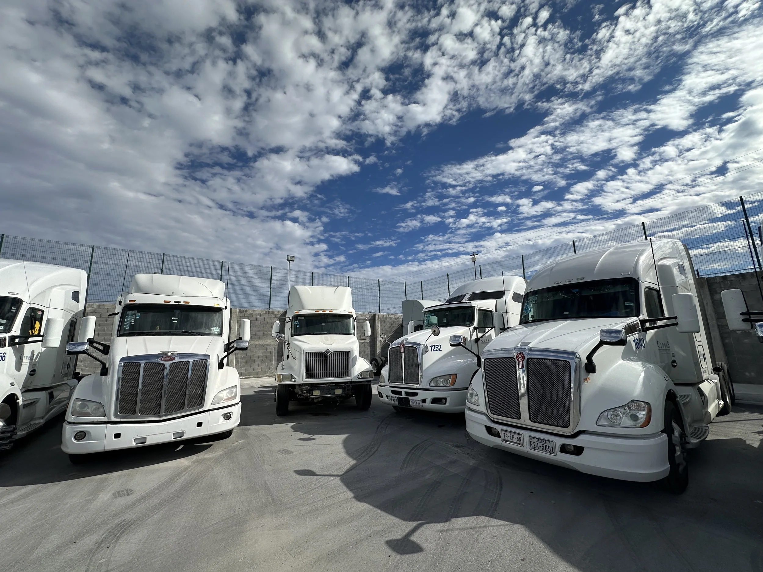 A parking lot filled with white semi-trucks parked in front of a high metal fence, with a partly cloudy sky overhead.