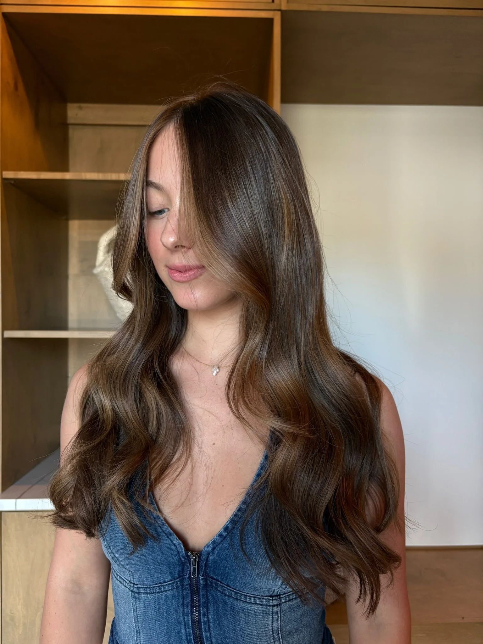 A woman with long, wavy brown hair and a side part, wearing a sleeveless denim dress with a zipper in the front, standing indoors in front of a wooden and beige background.