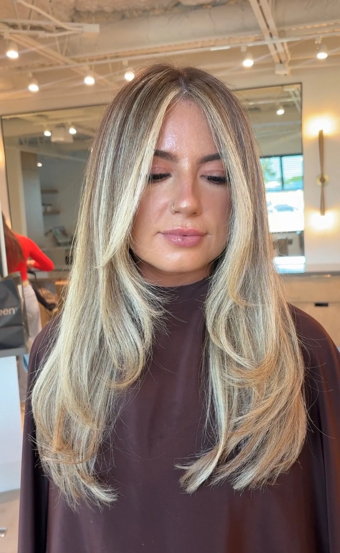 A woman with long, layered blonde hair and a nose ring in a salon, wearing a dark brown cape, with her eyes closed.
