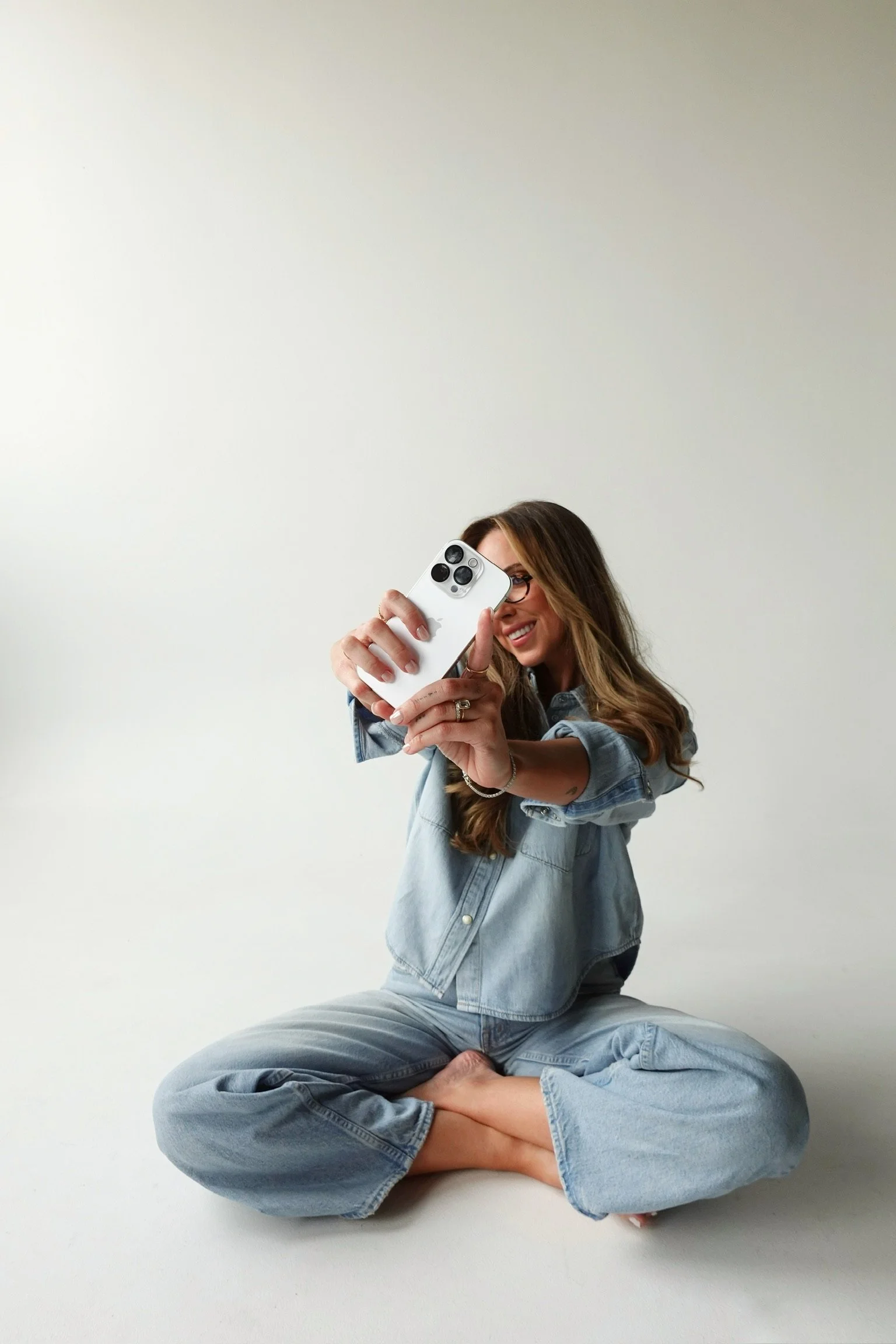 Woman with long brown hair and glasses sitting cross-legged while taking a selfie with her smartphone. She is wearing a denim jacket and pants, smiling at the camera against a plain, light-colored background.