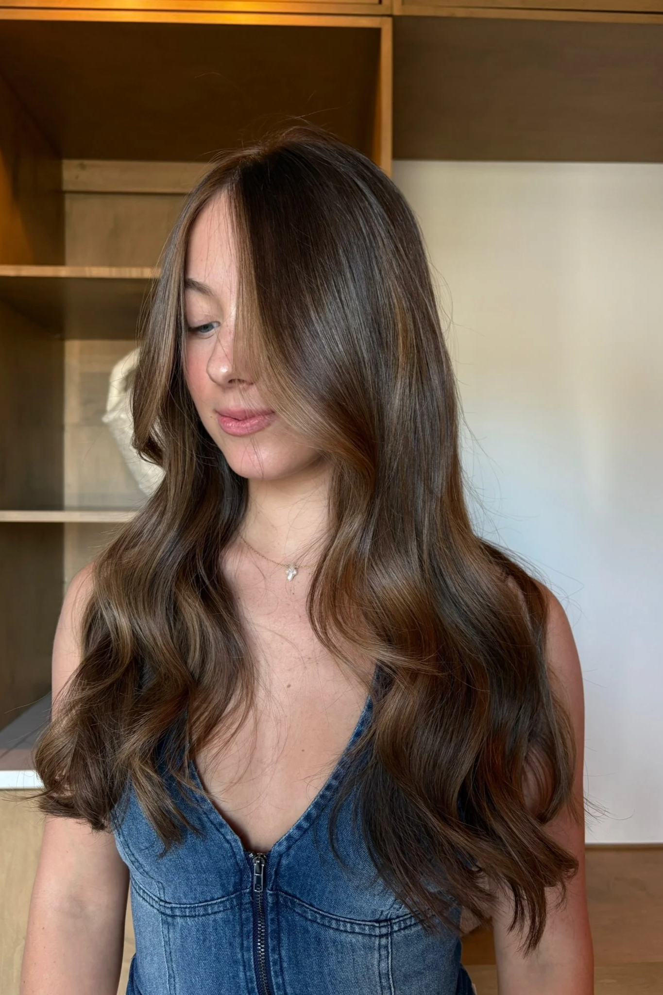 A woman with long, wavy brown hair and fair skin is looking down with her face partially covered by her hair. She is wearing a sleeveless blue denim top with a zipper in the front and a delicate necklace. The background features a wooden shelving unit and beige walls.