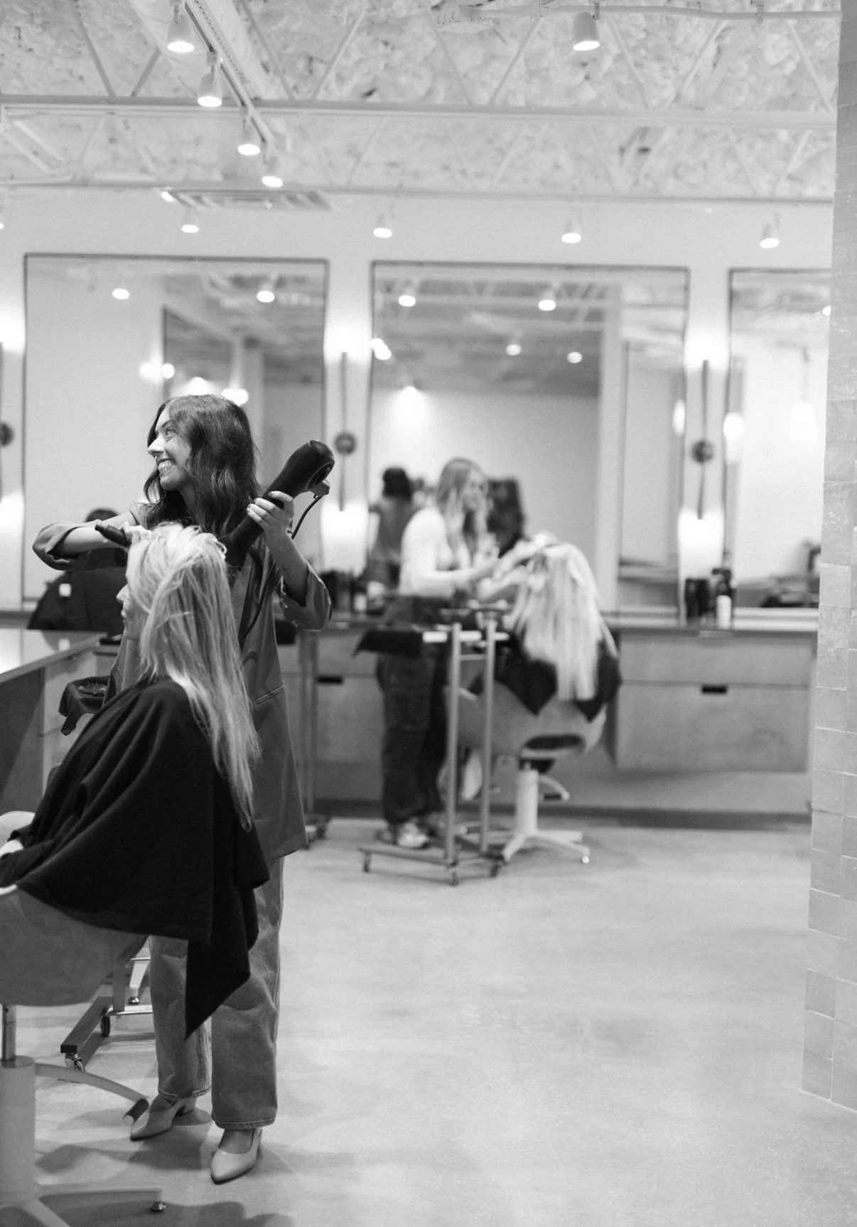 A hairstylist drying a woman's hair in a salon. The woman is seated with a cape, and others are visible in the background working or waiting.