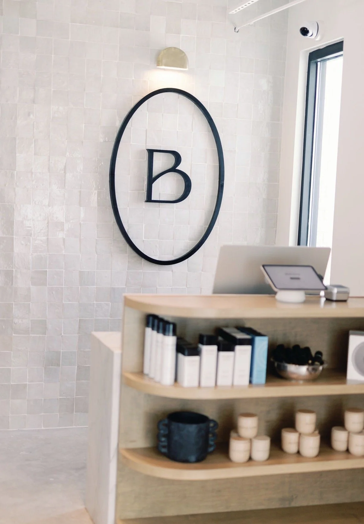 Reception area with a large black oval frame and letter 'B' on a light beige tiled wall, a small wooden shelf with books, a black pot, and cream-colored cups, and a receipt or sign on a countertop with a computer and a window in the background.