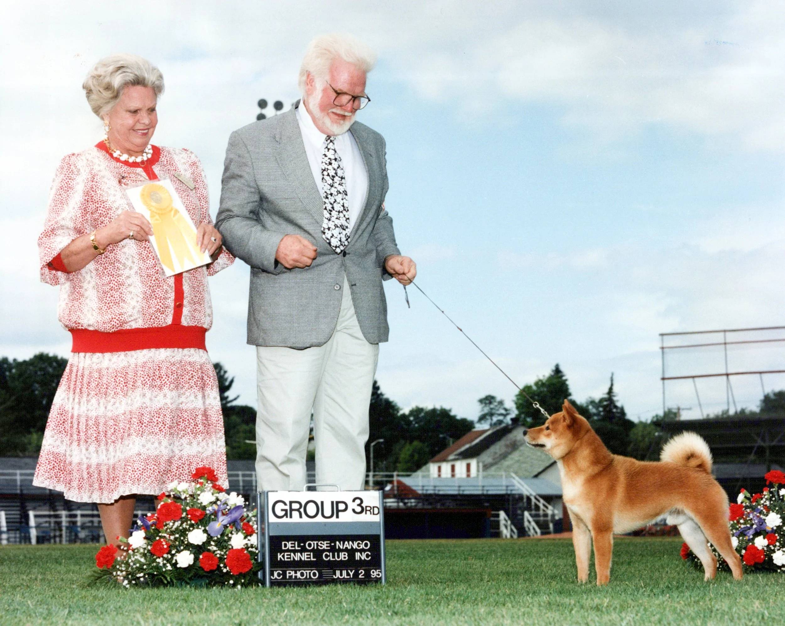 An elderly woman and man stand on a field at a dog show. The woman holds a ribbon and has a display of flowers at her side. The man is holding a leash attached to a medium-sized, brown and white dog. There is a sign indicating they are in the third g