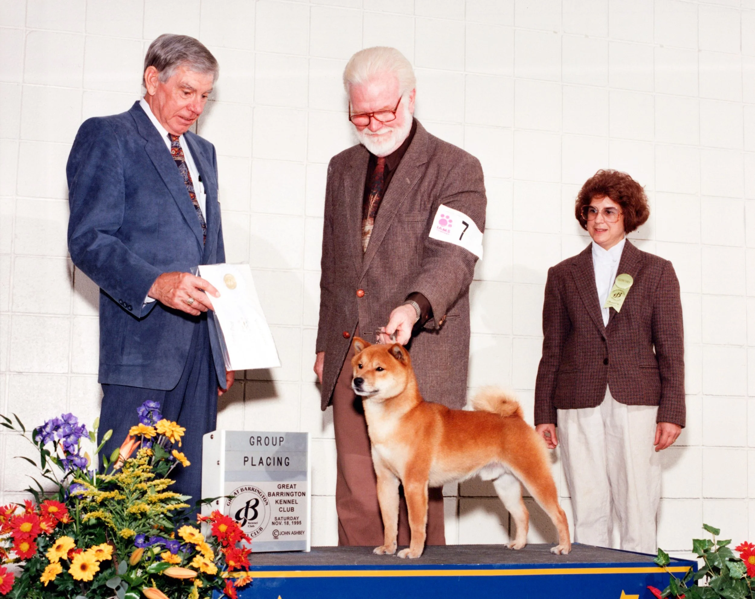 Three people in formal attire and a Shiba Inu dog pose on a stage at a dog show with flowers and a sign. The man on the left is holding a certificate, the man in the center is looking down at the dog, and the woman on the right is smiling with a ribb