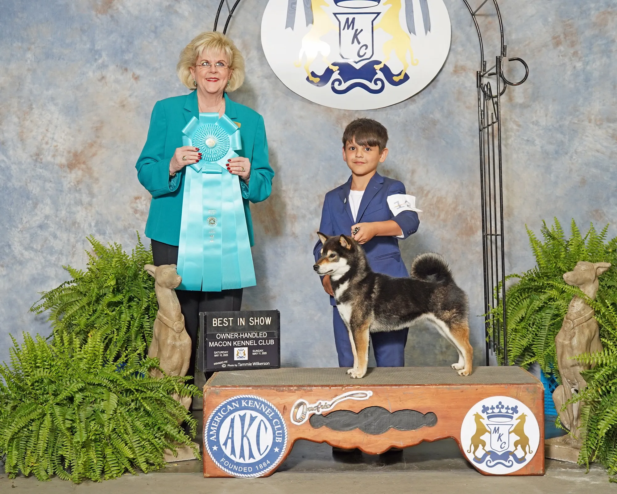 An award ceremony at the Macon Kennel Club, featuring a woman and a young boy with a Siberian Husky, along with plants and a 'Best in Show' sign.