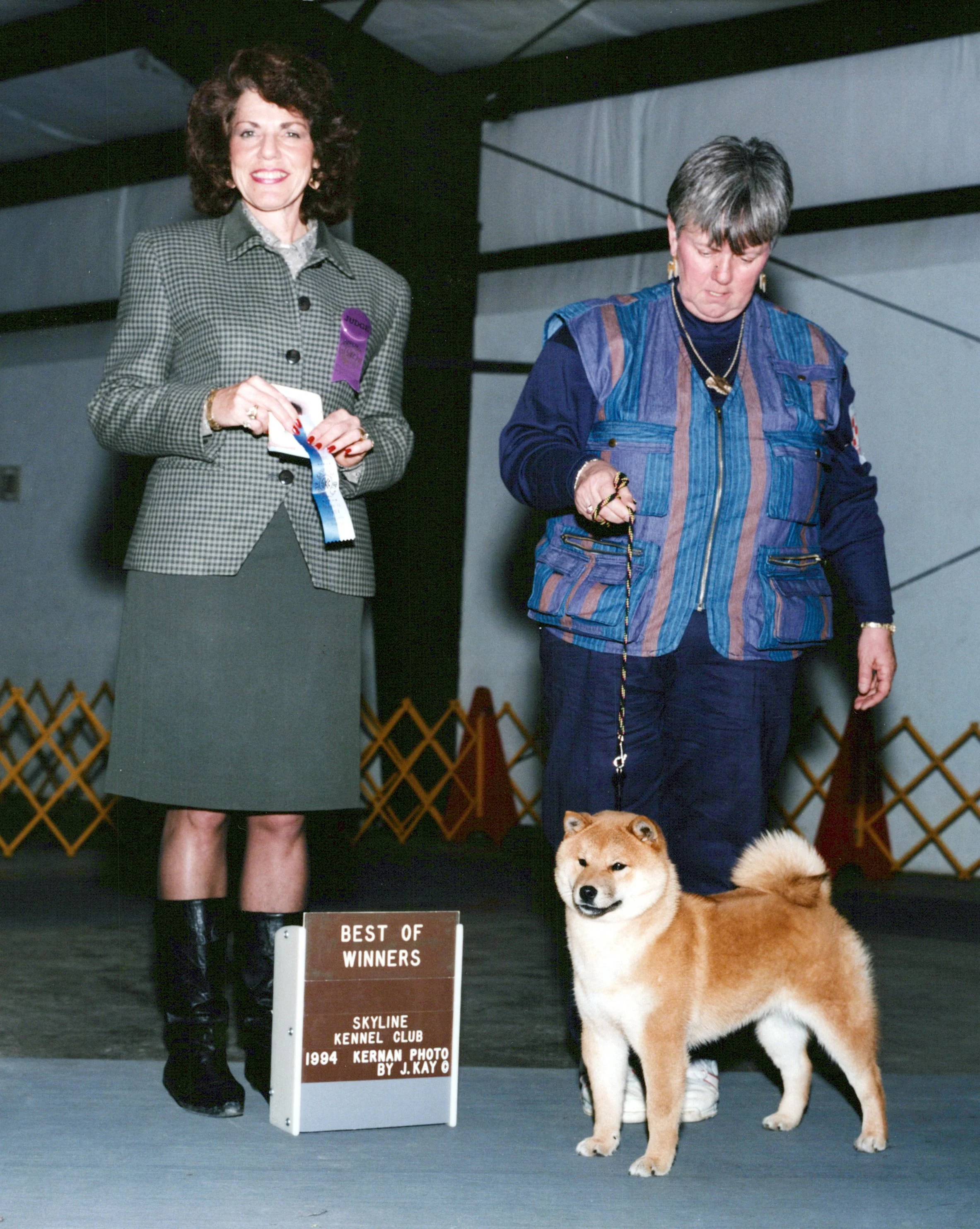 A woman in a checkered blazer holding a ribbon stands next to a woman with a short hairstyle wearing a denim jacket. Two Shiba Inu dogs are in the foreground, one with a thick, fluffy coat. The scene appears to be at a dog show, with a sign reading "