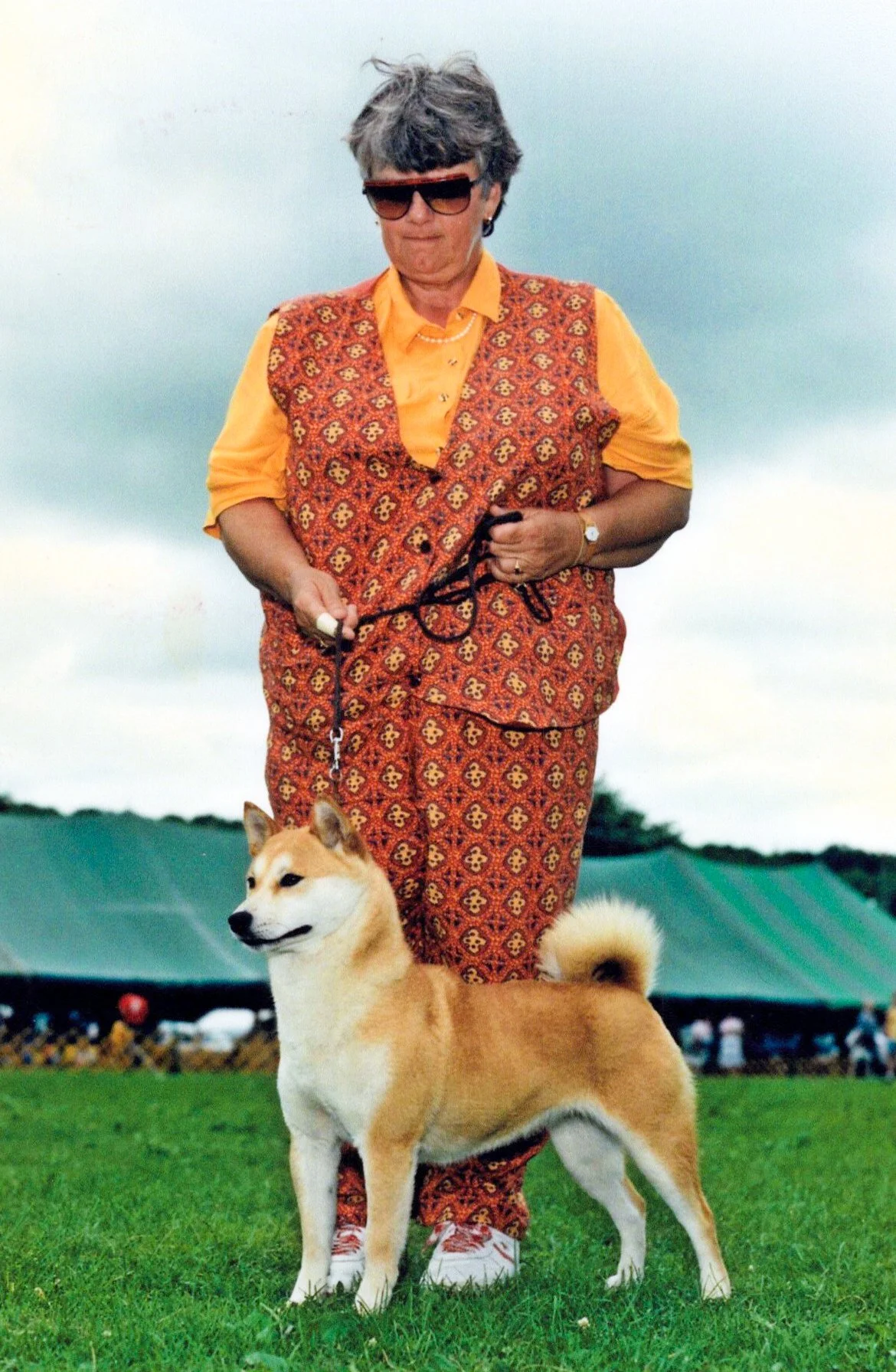 An elderly woman wearing sunglasses, a yellow shirt, and patterned vest and pants walks a dog on a leash in a park. The dog is a fluffy, tan and white breed, standing on green grass with a large tent and trees in the background.