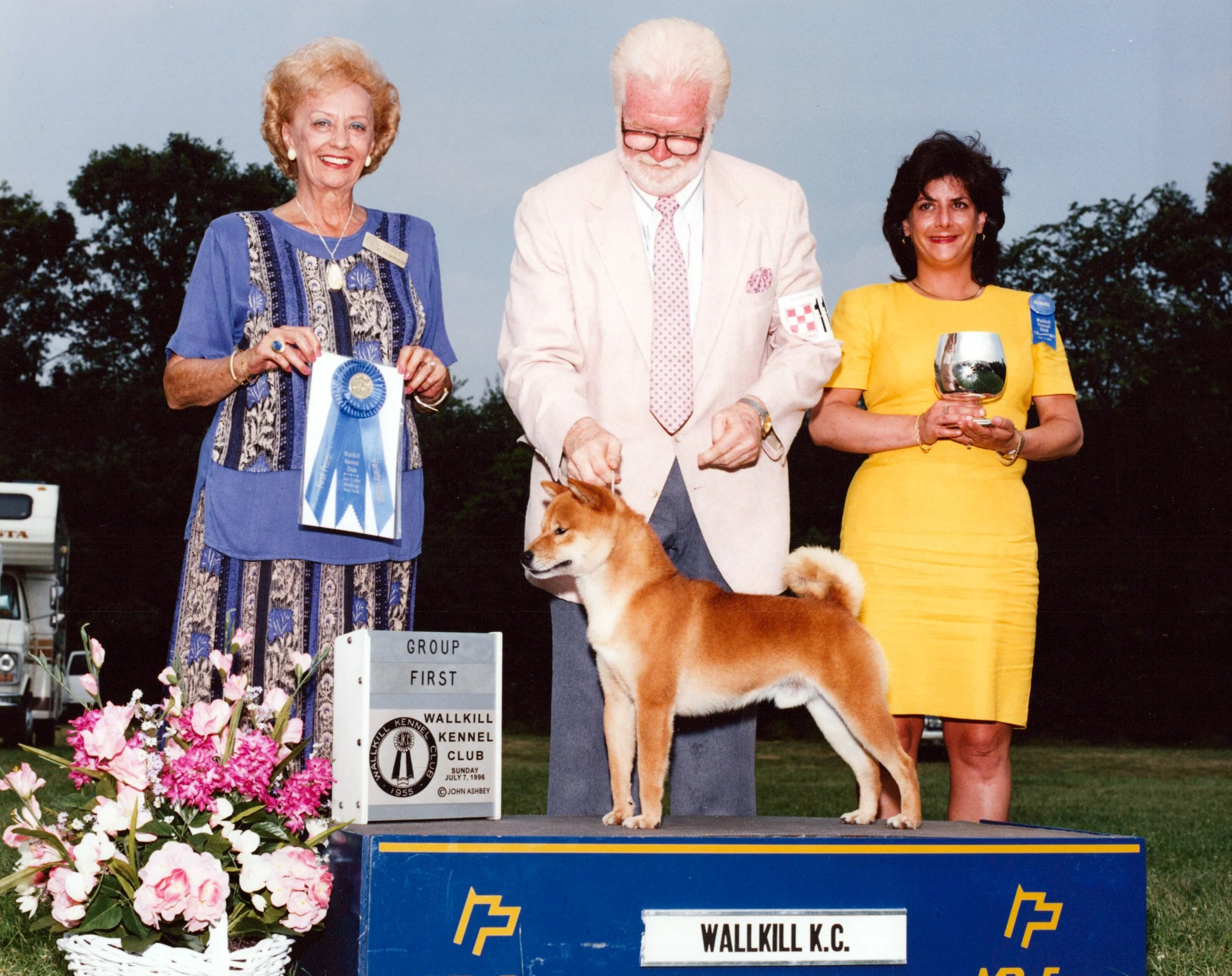 Three people standing outdoors at a dog show, with flowers in a basket in the foreground and a sign that reads 'Wallkill K.C.' in the background. One woman wears a blue dress and holds a blue ribbon, a man in the middle with white hair, glasses, and a beige suit, and another woman in a yellow dress holding a silver bowl. The dog, a Shiba Inu, is standing on the platform in front of them.