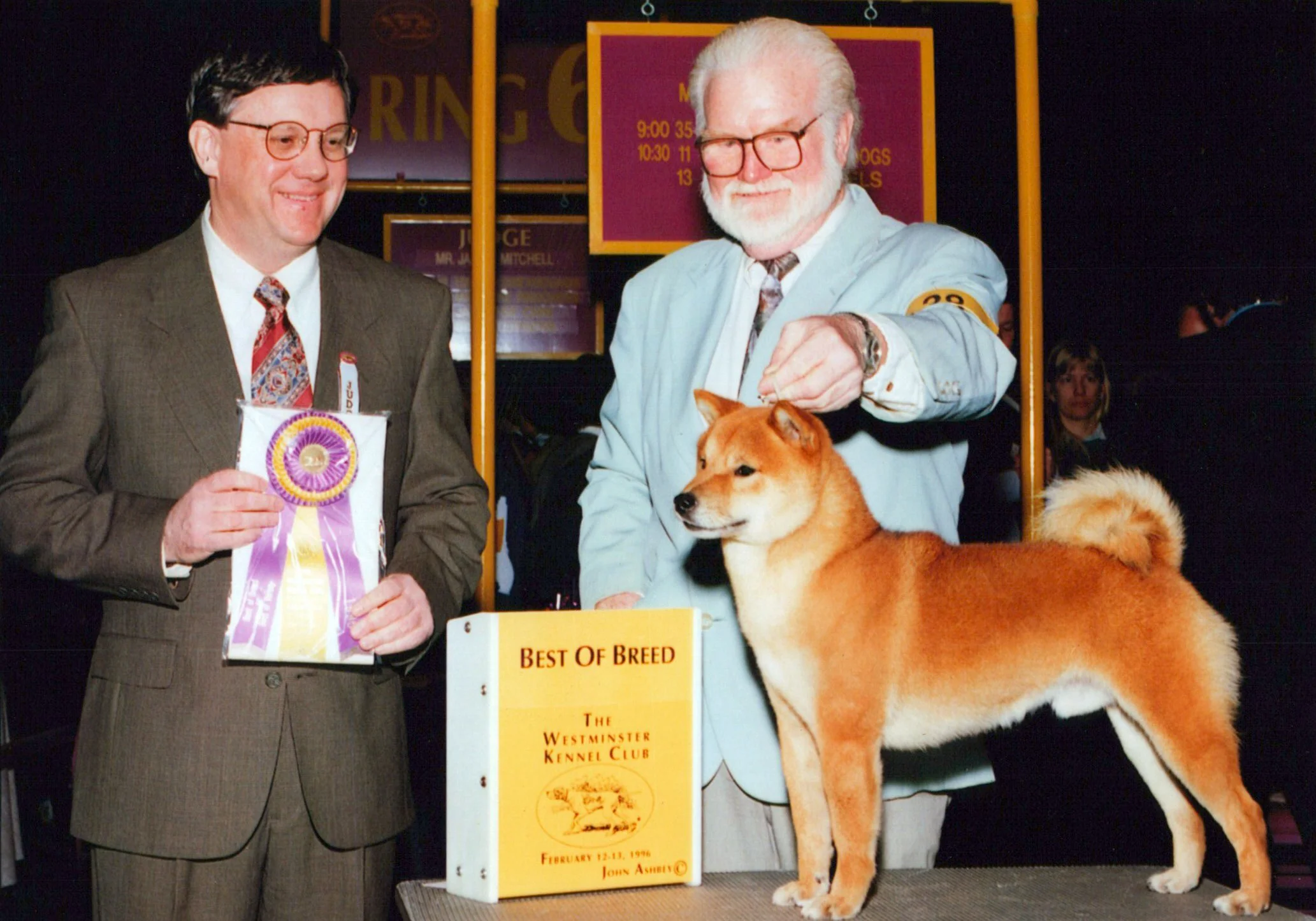 Two men in suits and glasses standing next to a Shiba Inu dog and a banner at a dog show. One man holds a ribbon, and there is a sign that reads 'Best of Breed' with details about the event.