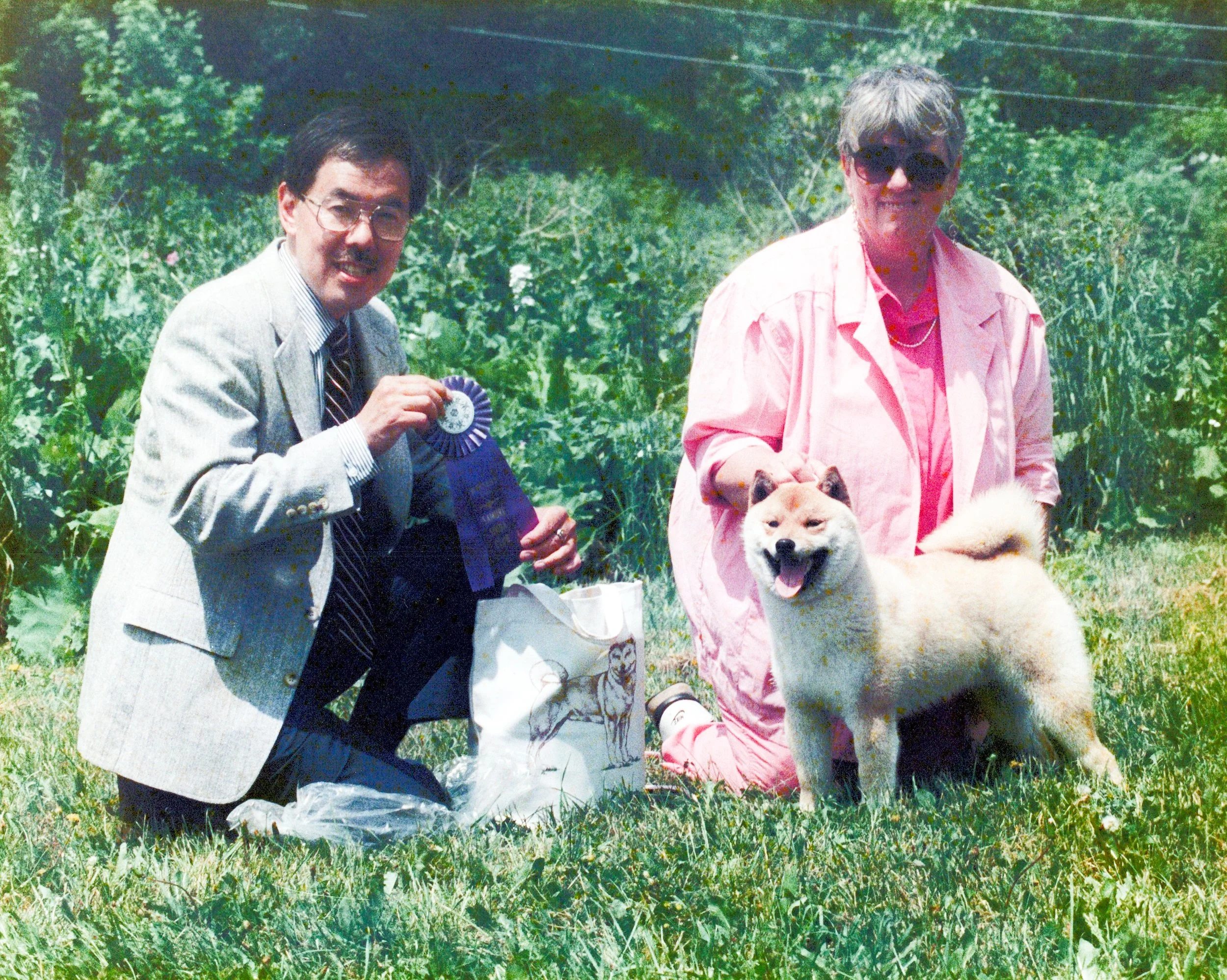 Two men with dogs outdoors on a grassy field with trees, one holding a rosette ribbon, the other with a sitting dog.