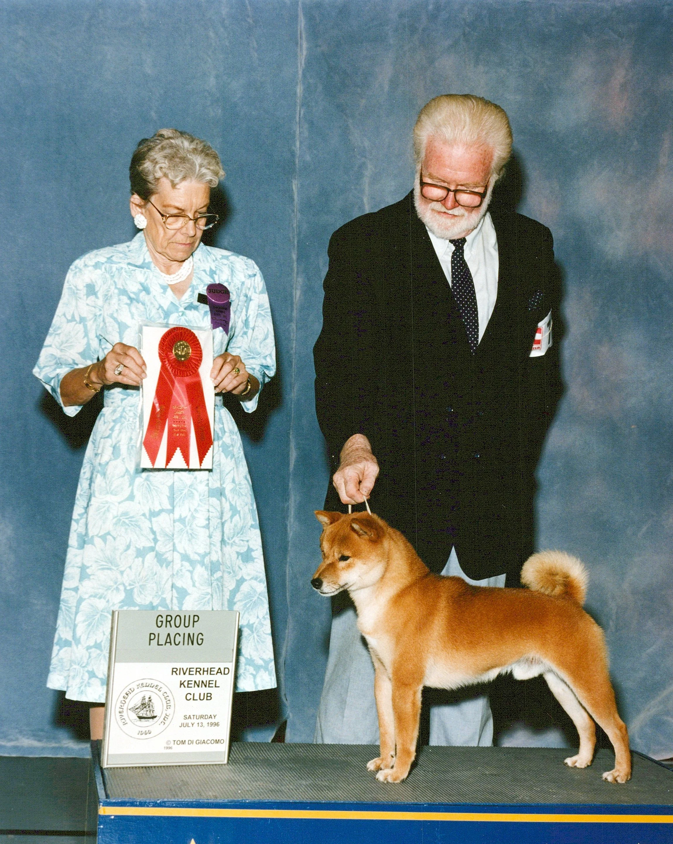 A woman holding a ribbon and a man standing next to a Shiba Inu dog at a dog show, with a sign indicating the group placing and event details.