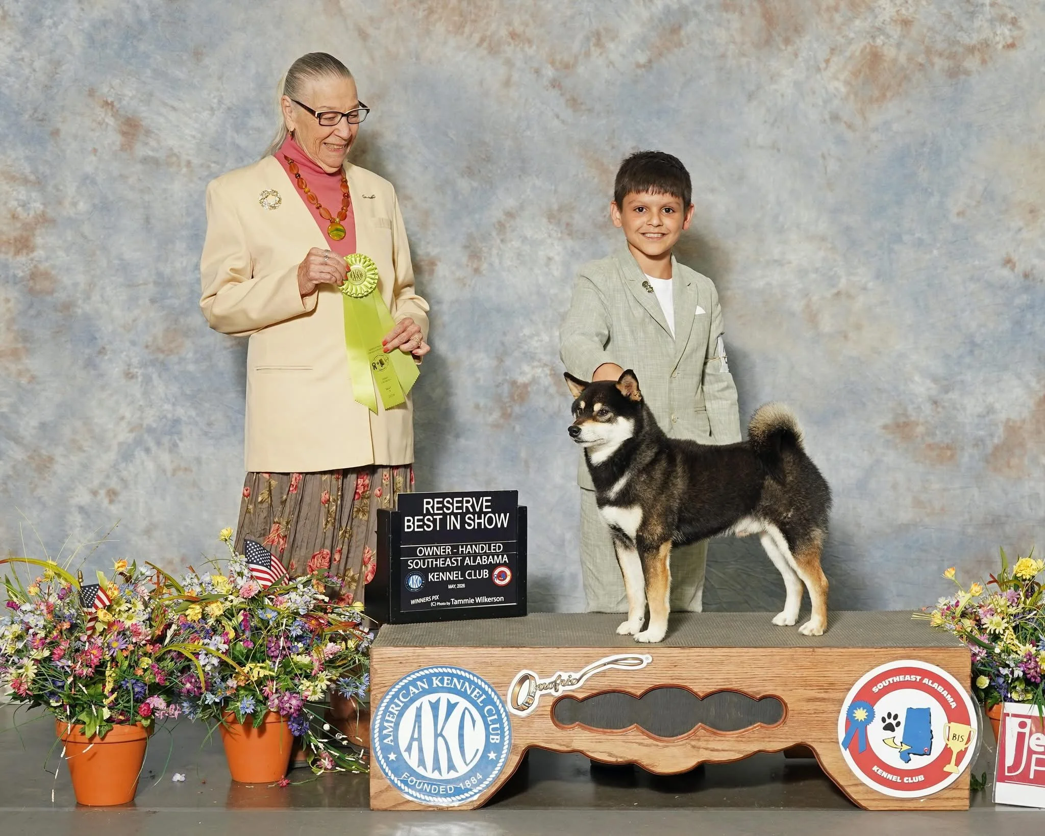 A woman and a young boy standing on a stage behind a black sign with white lettering, holding a ribbon. The woman is wearing glasses, a cream-colored blazer, and a pink turtleneck, holding a yellow ribbon. The boy is dressed in a light gray suit, smiling, with a Siberian Husky dog in front of him. The stage is decorated with floral arrangements and badges, with a backdrop of a cloudy sky pattern.