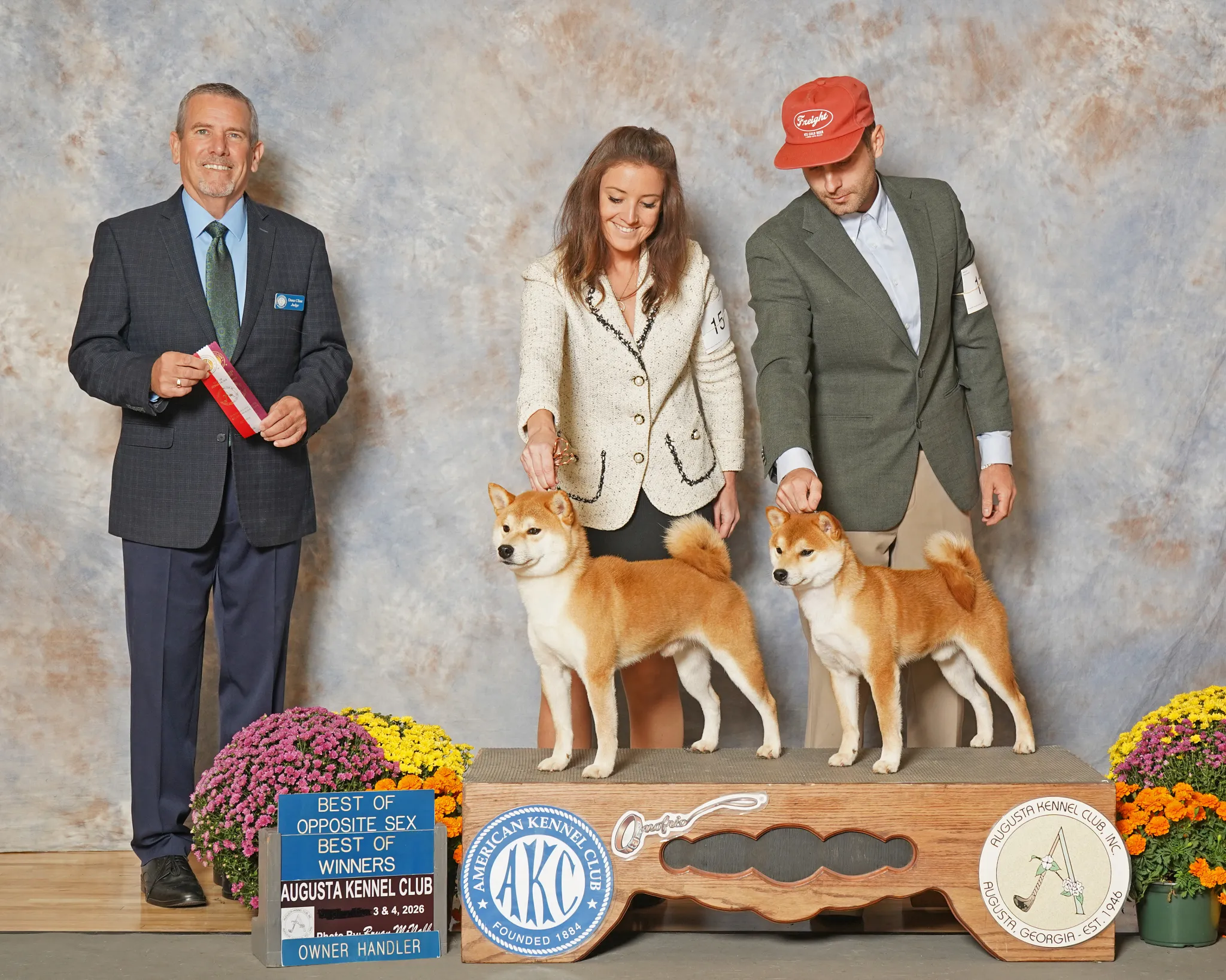 Three people standing behind a row of three Shiba Inu dogs at an award event, with flowers and signs indicating the event's details and awards on display.