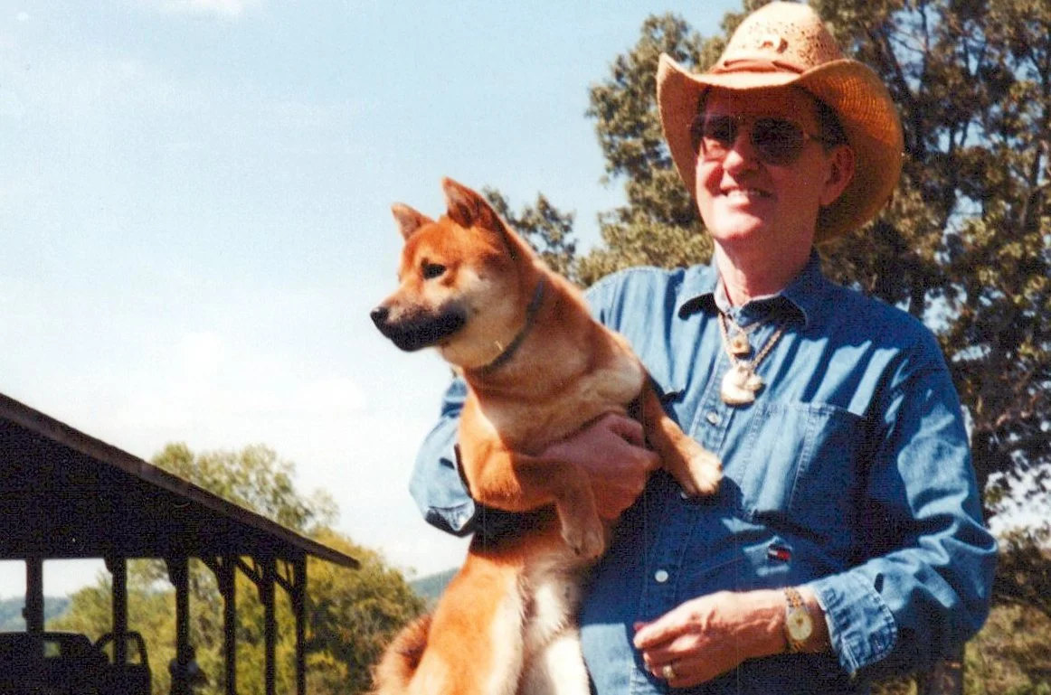 A woman wearing a cowboy hat, sunglasses, and a denim shirt holding a tan and black dog outdoors with trees and a blue sky in the background.