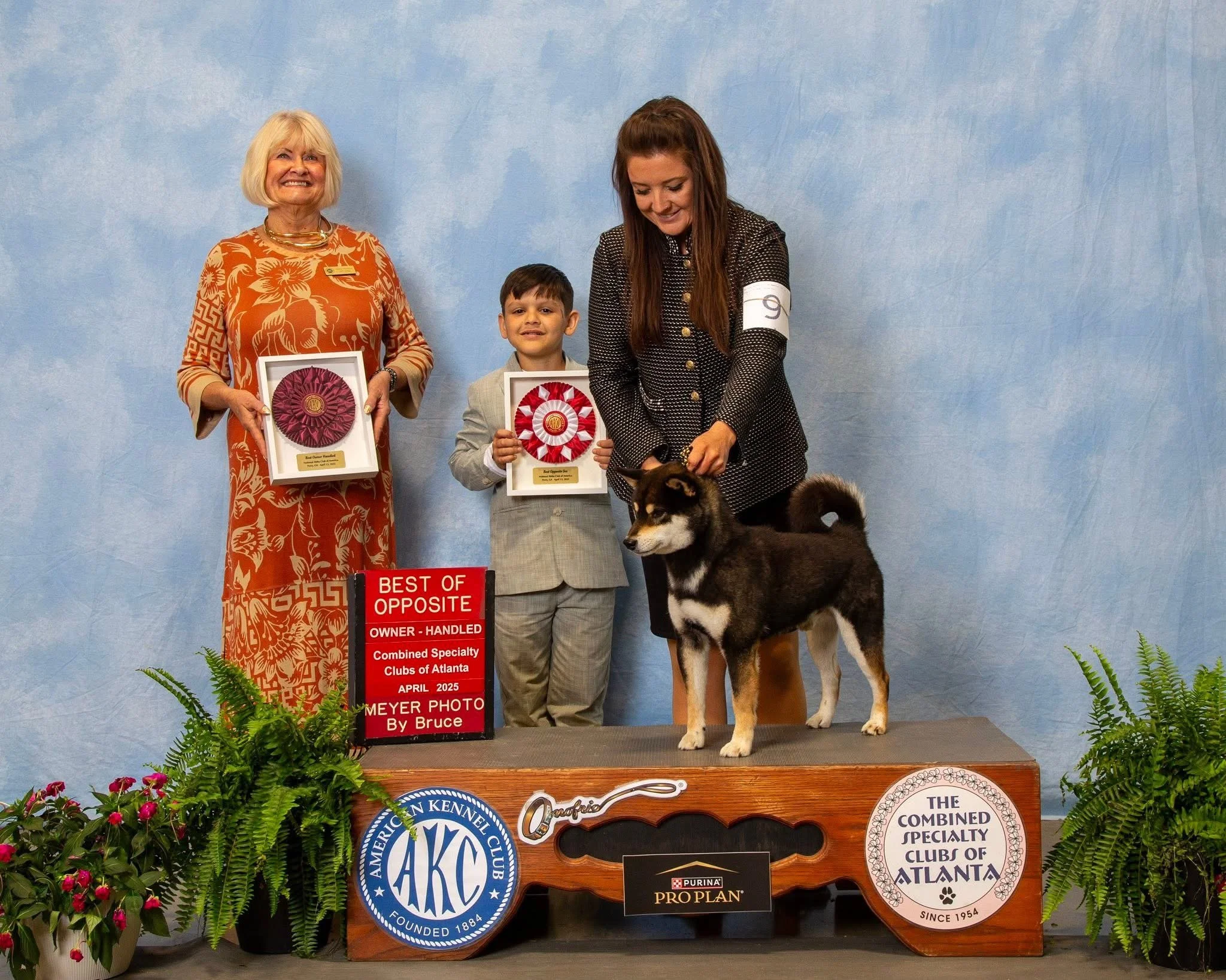 A woman, child, and woman with a dog at a dog show, holding award ribbons and standing on a podium. The woman on the left is wearing an orange patterned dress, the woman in the middle is in a gray suit, and the woman on the right is in a black and white outfit handling a black and white dog. The background is light blue with potted plants and a red sign that says "Best Opposite" and other text.
