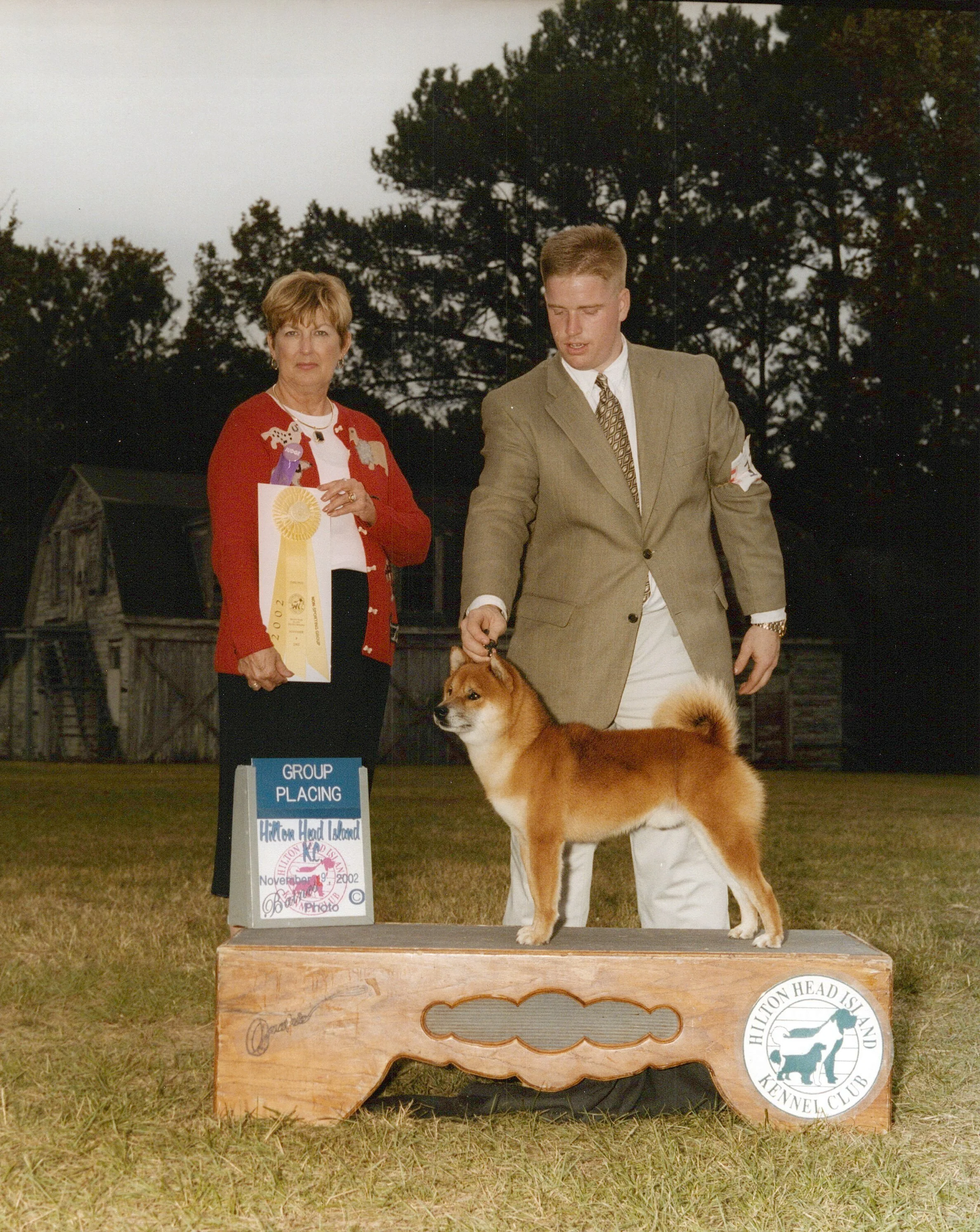 Dog and handler at a dog show, with a ribbon and group placings sign, on a grassy field during daytime.