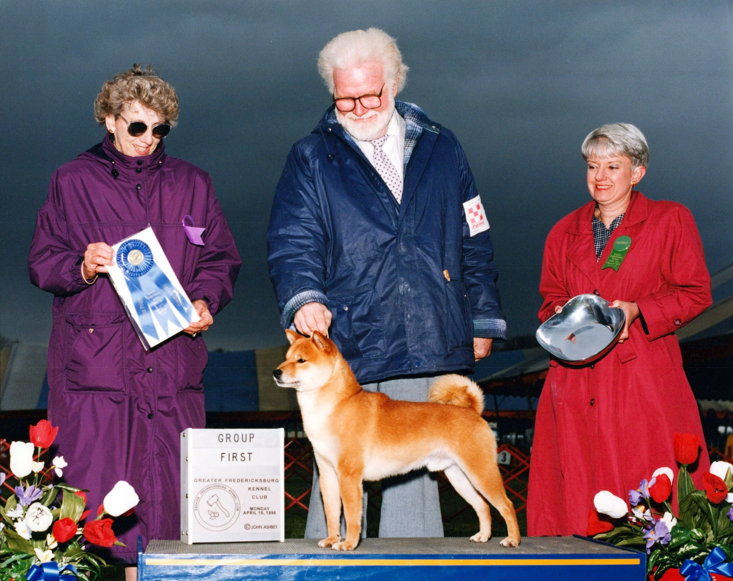 A group of three elderly people, two women and one man, standing behind a golden retriever dog on a platform at a dog show. The women are wearing purple and red jackets, both with sunglasses, and the man is dressed in a blue jacket with a plaid shirt underneath. They are smiling and looking down at the dog, which is standing on the platform. There are flowers and a sign indicating the dog won first place in the group, with details about the event and date.