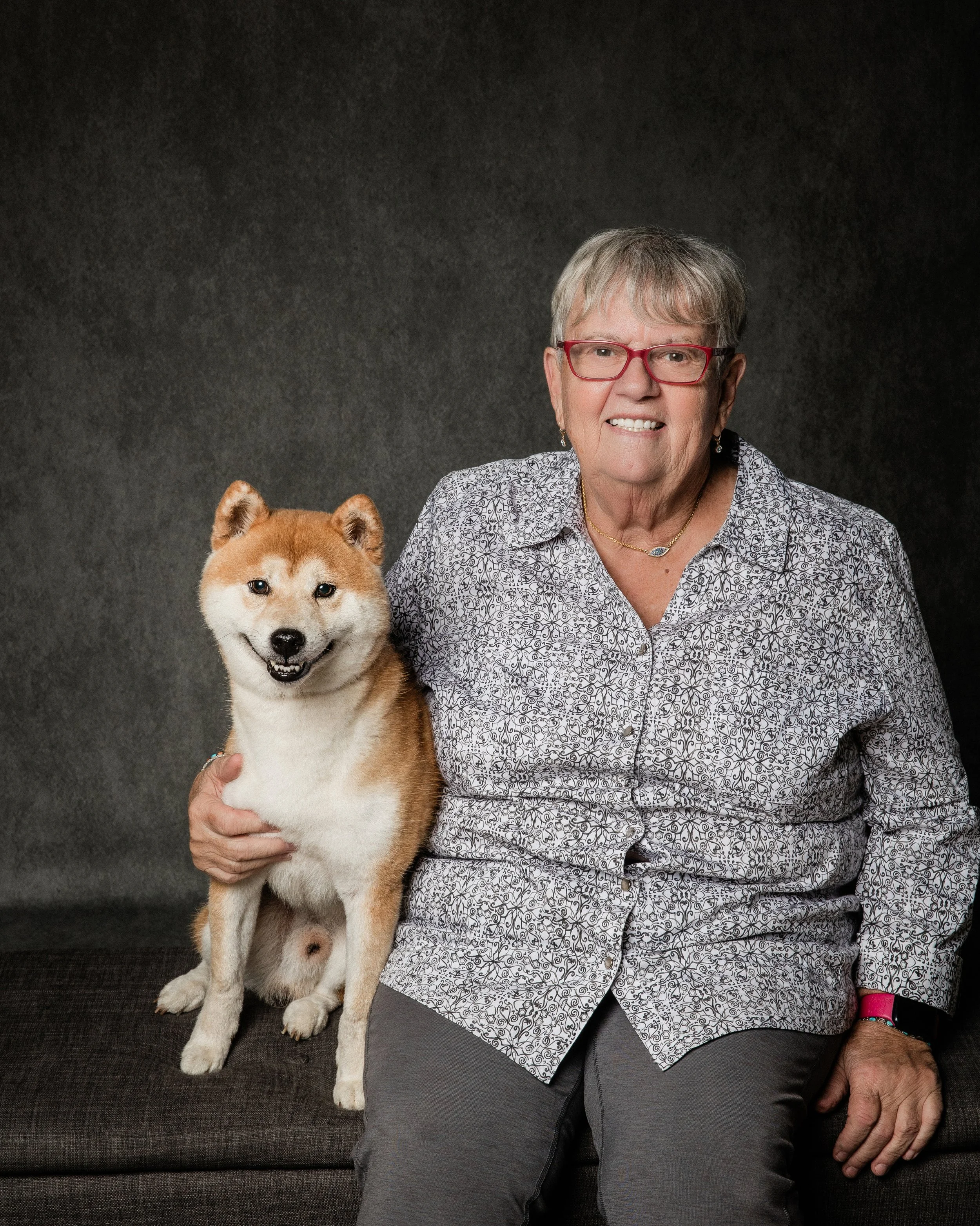 Senior woman with short gray hair, red glasses, and gold jewelry, sitting on a dark gray couch with a Shiba Inu puppy, both smiling, against a gray background.