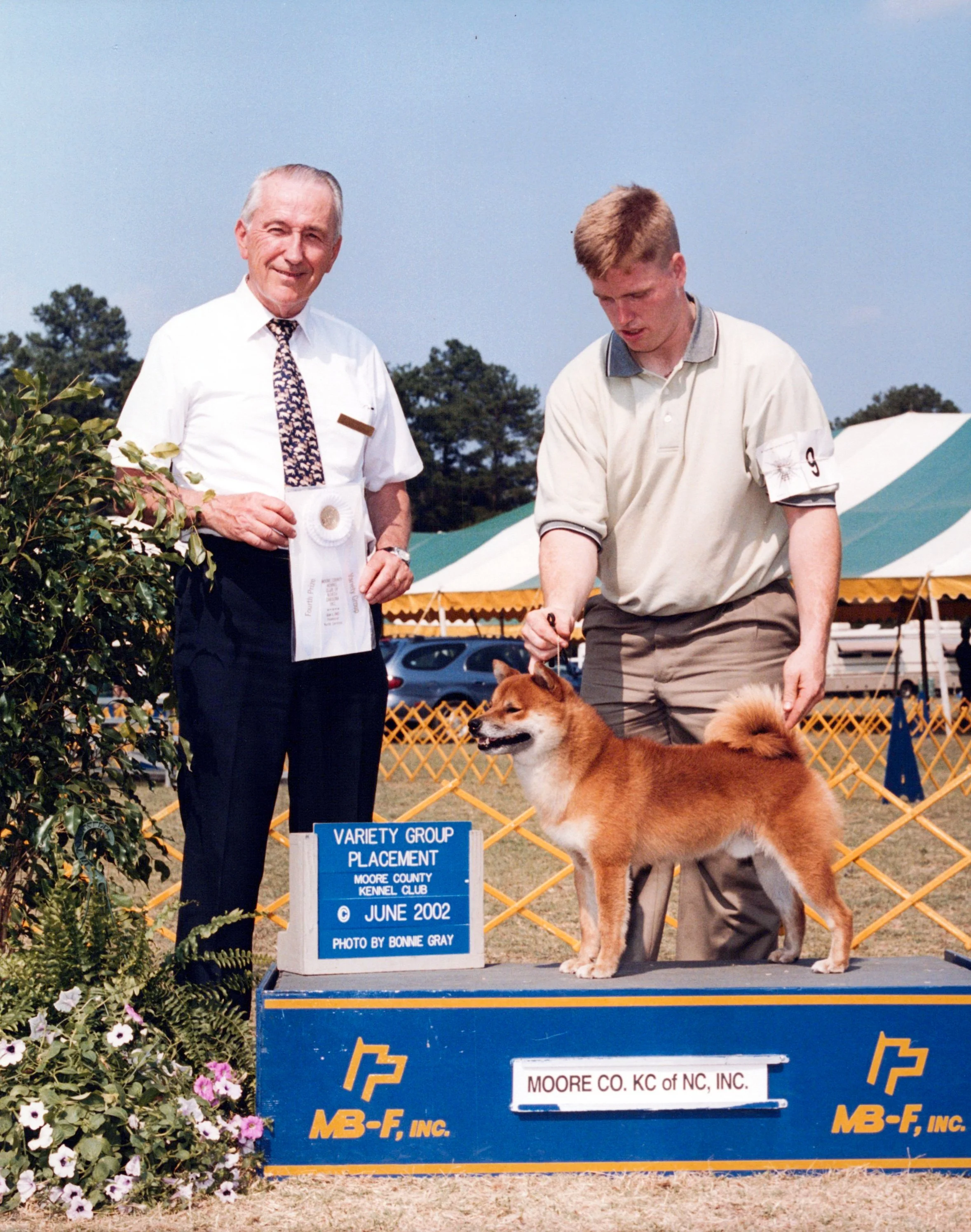 A man presenting a brown and white dog at a dog show, with a judge holding a ribbon. The event is organized by Moore County Kennel Club, held in June 2002, with a sign indicating the "Variety Group Placement." The background features tents and trees,
