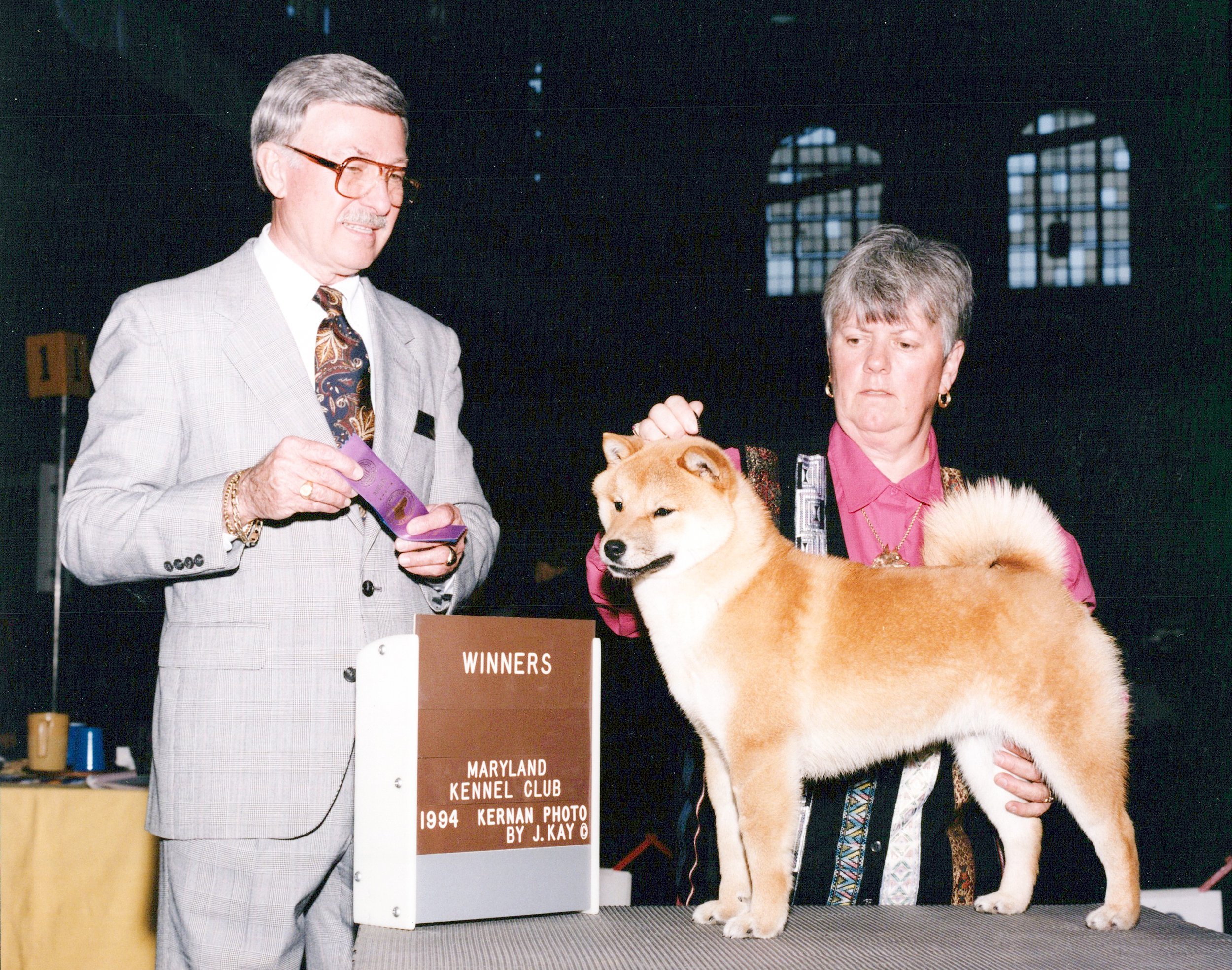 A man in a light gray plaid suit with glasses and a woman in a pink jacket holding a Shiba Inu dog at a dog show, with a sign indicating winners from Maryland at the Maryland Kennel Club in 1994.