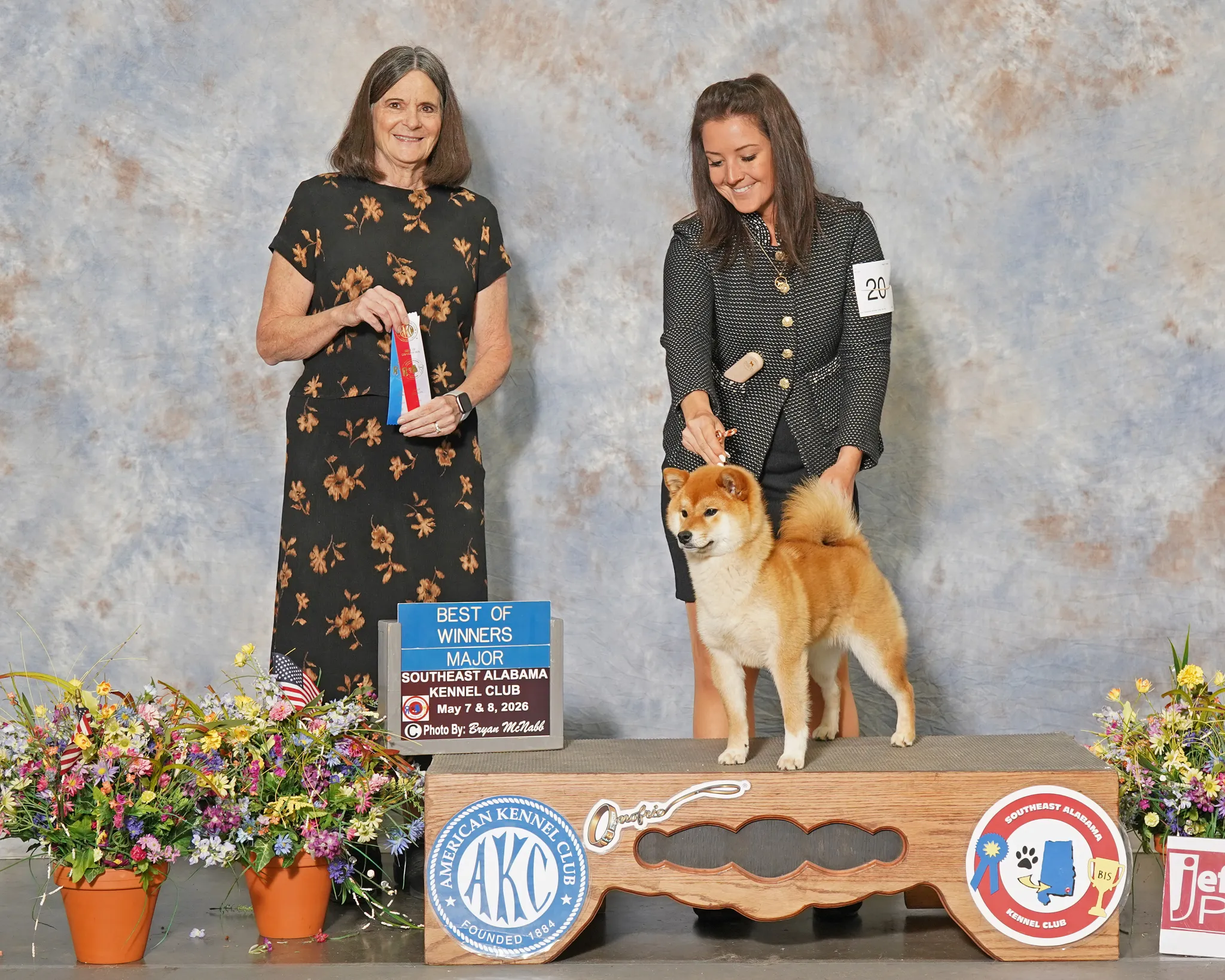 Two women with a Shiba Inu dog on an awards podium at a dog show. The woman on the left is wearing a black dress with orange flowers and holding a ribbon. The woman on the right is wearing a black and white polka dot jacket, holding the dog's leash. The podium has a sign that says "Best of Winners Major Southeast Alabama Kennel Club May 7 & 8, 2026" and various logos and awards. There are colorful flowers in pots around the podium.