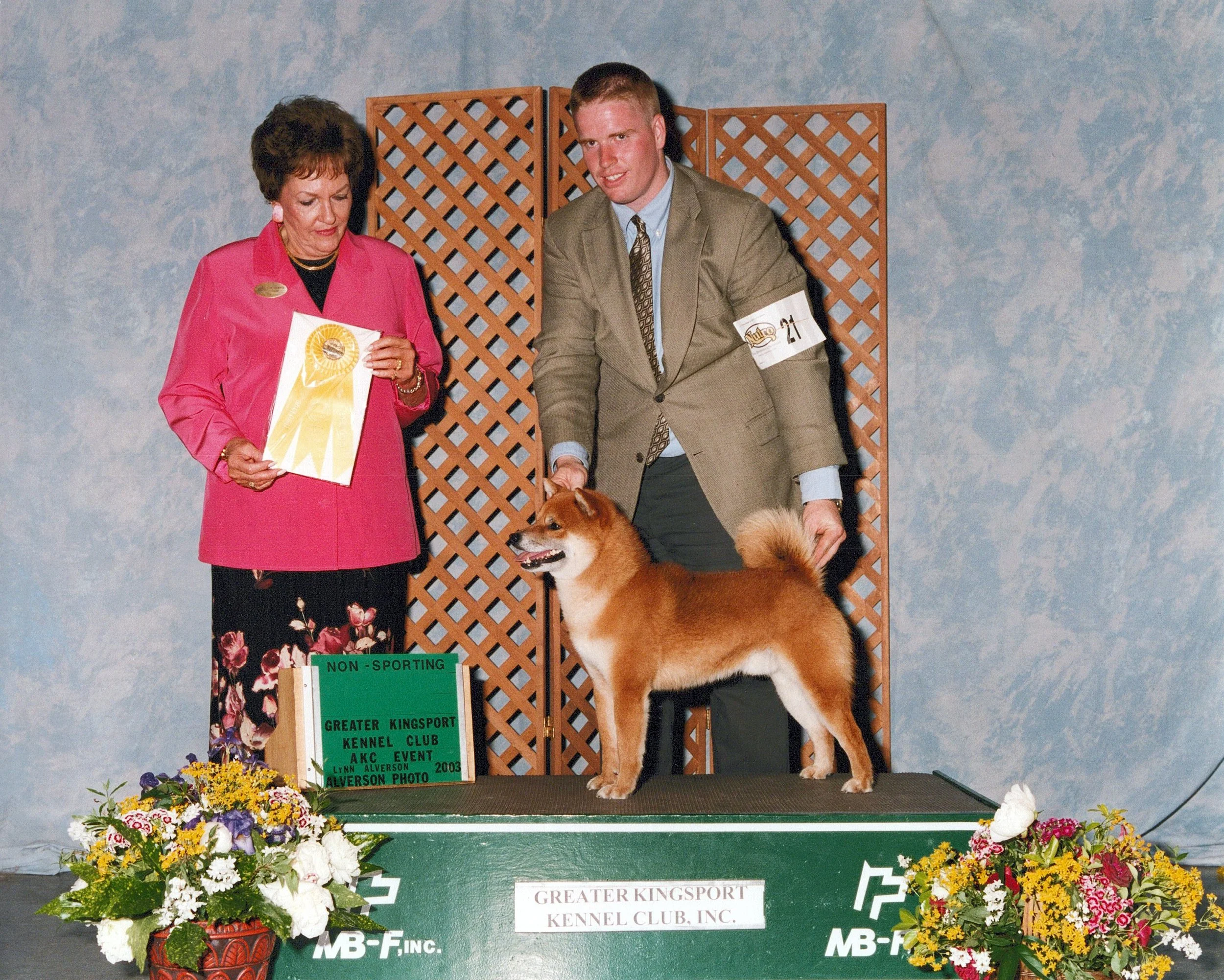 A young man in a tan suit and a woman in a pink blazer are standing on a platform with a dog. The woman is looking down at a ribbon award, and the young man is looking at the camera with a smile. The platform has a sign that reads: 'Greater Kingsport