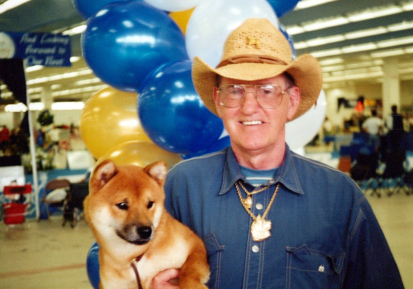 A man with glasses, wearing a cowboy hat and denim shirt, is holding a puppy at an indoor event with blue, white, and gold balloons in the background.