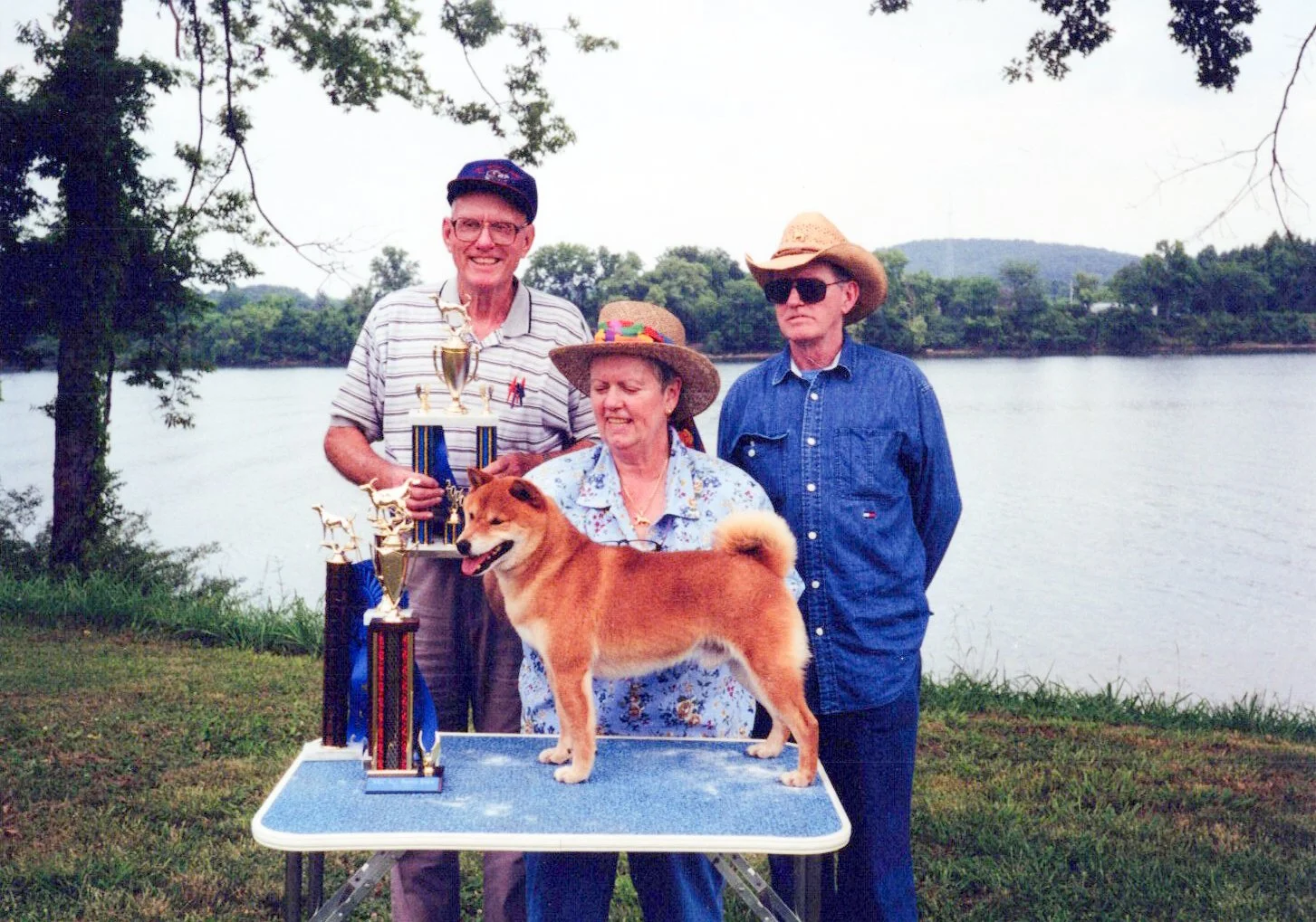 A group of three adults and a dog outdoors near a body of water, with trophies displayed on a table, celebrating a dog show.
